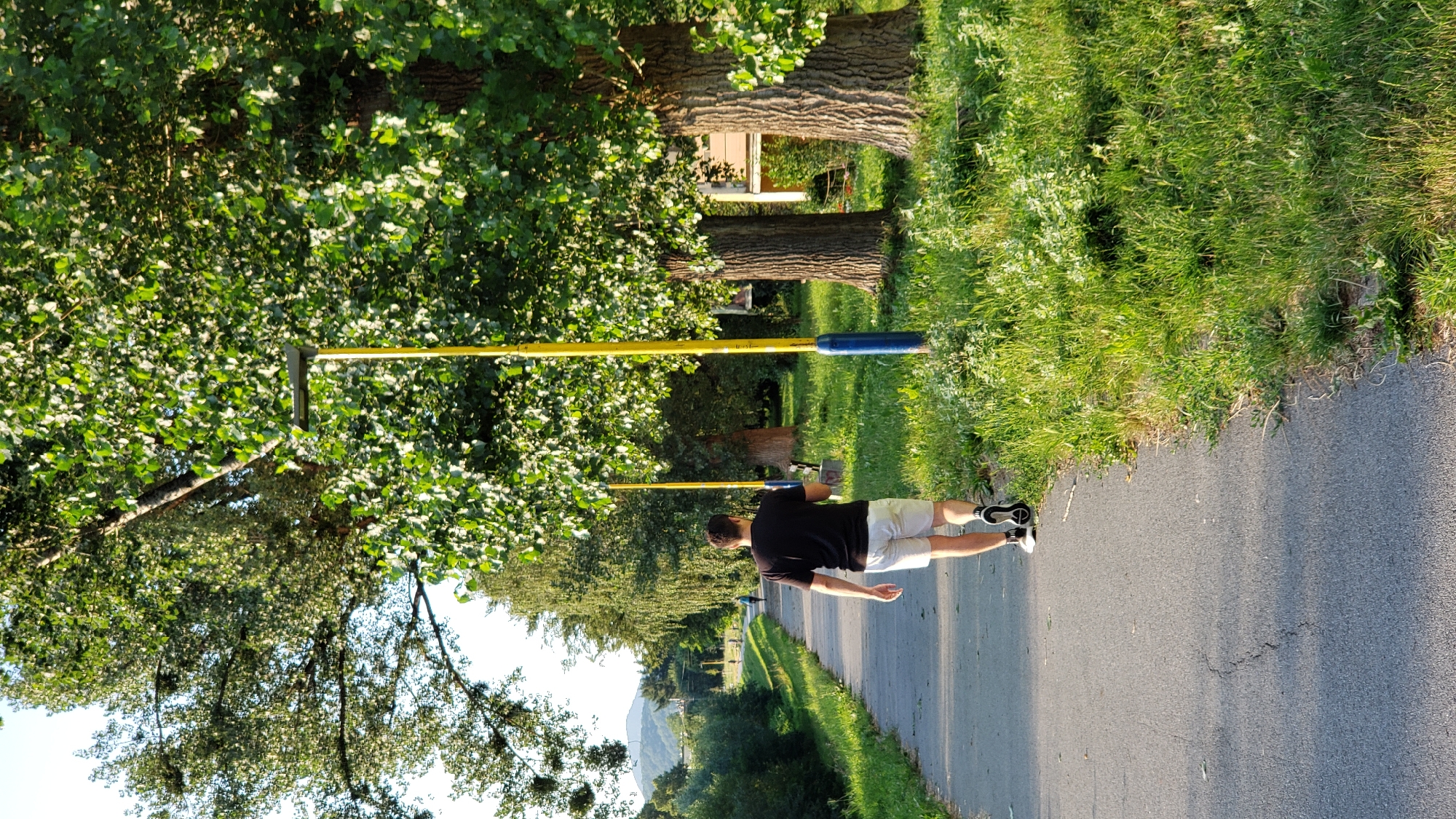 The image shows a person walking along a paved path in Prešov, Slovakia. The person is wearing a black t-shirt and light-colored shorts with athletic shoes. They are walking away from the camera, towards a line of trees. The path is bordered by green grass and more trees. The sun appears to be low in the sky, casting long shadows, suggesting it is either early morning or late afternoon. The overall scene is one of a pleasant outdoor walk, likely during warm weather. The environment is natural and serene, with a focus on the greenery and the path.