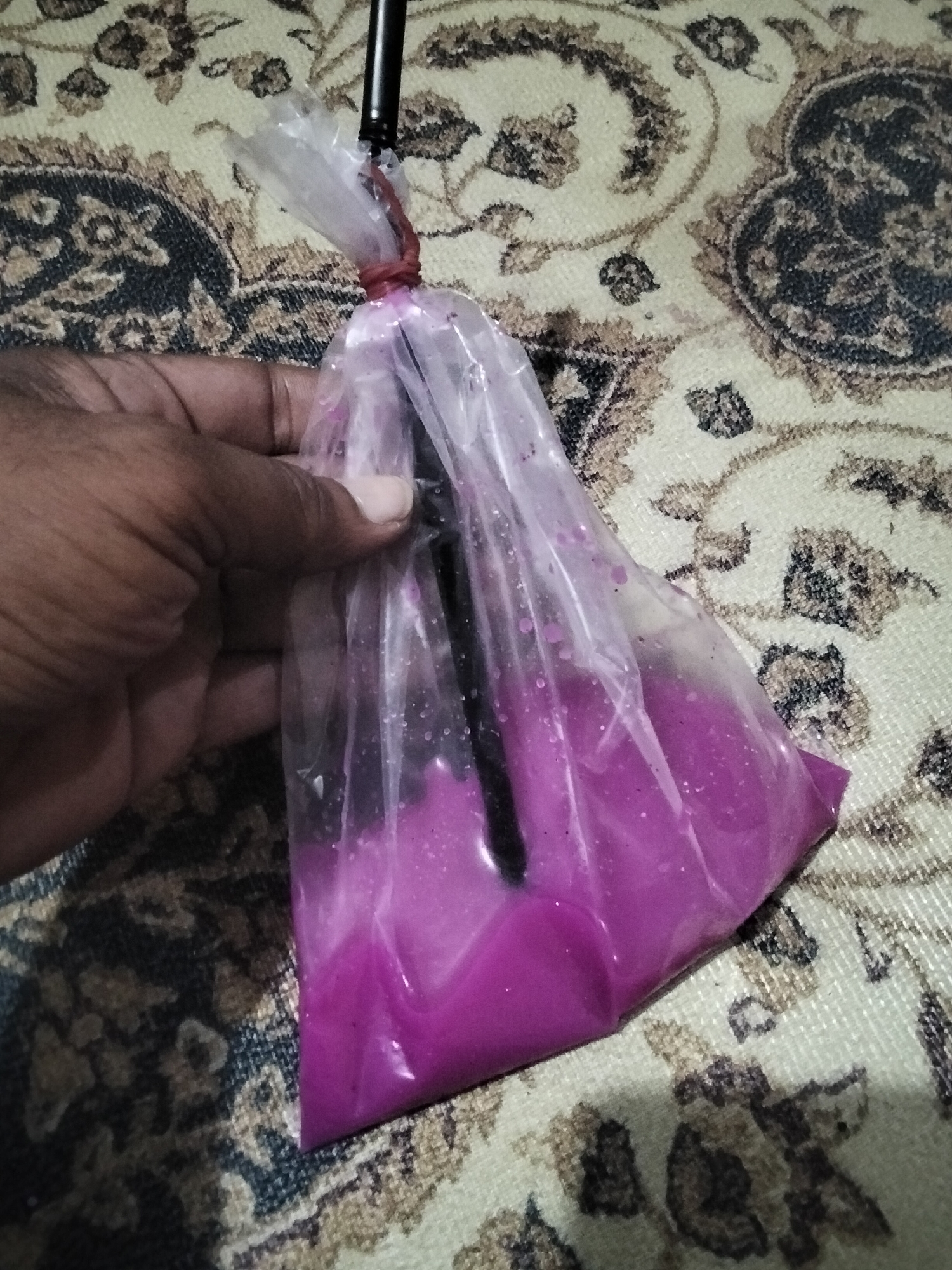 A hand holds a clear plastic bag containing a bright purple liquid and a black straw, with a red rubber band securing the top of the bag around the straw. The bag rests on a patterned surface of cream, brown, and black. The location, Lhokseumawe, Indonesia, is unknown based solely on the visual information.