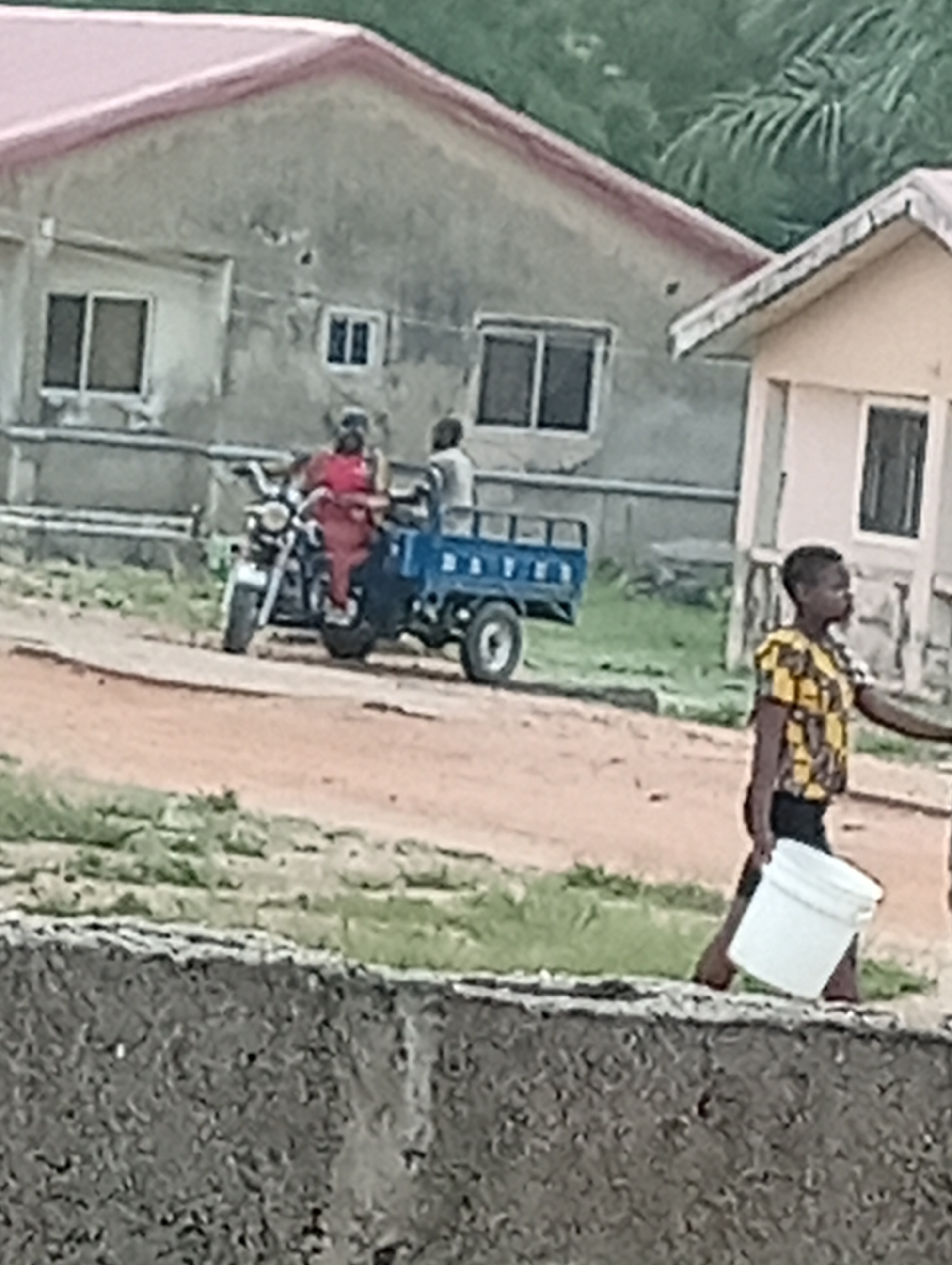 In Adankolo, Nigeria, a boy carrying a white bucket walks towards the camera. Behind him, a three-wheeled motorcycle with two people is parked on a dirt road in front of houses. The houses have multiple windows. The foreground shows a gray concrete structure. The sky is overcast.