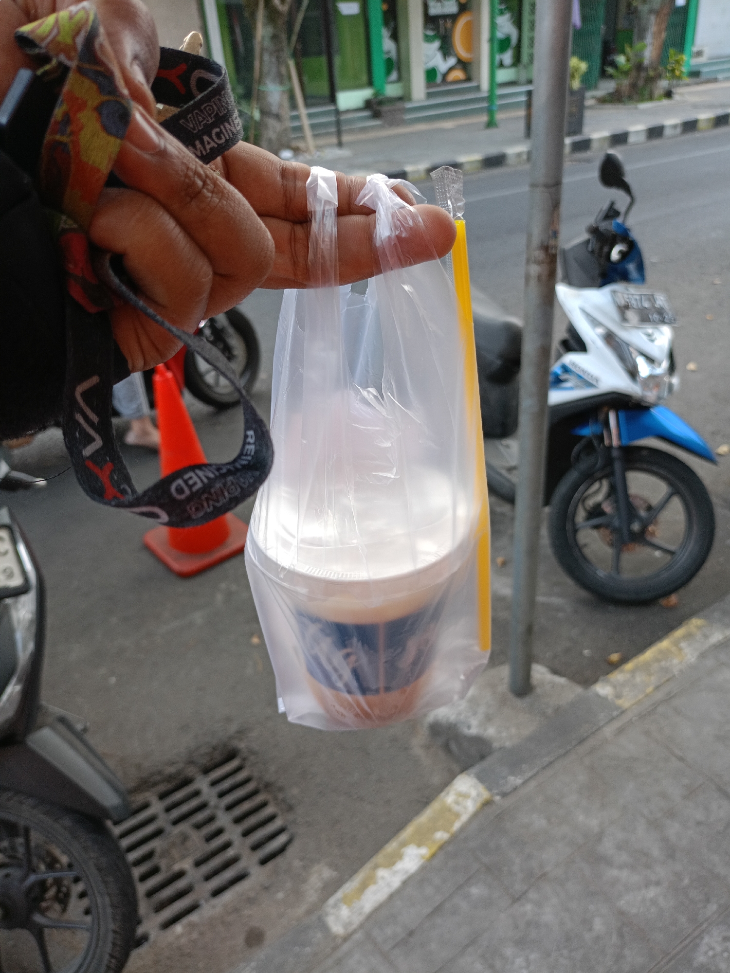 The image captures a close-up view of a person's hand holding a plastic bag containing a beverage and a straw. The bag is transparent, and the drink appears to be a light brown color, possibly coffee or tea. A yellow straw is visible sticking out of the bag. The person's hand has several rings and is holding a dark lanyard with colorful patterns. In the background, a street scene is visible, with a white and blue motorcycle parked on the right side of the frame. A bright orange traffic cone is also present on the left. The environment suggests an outdoor setting in a city, potentially in Batu, Indonesia, given the details of the motorcycle's license plate which reads "AG 5874 JK" and the general urban landscape. The pavement has a metal grate, indicating a drainage system. The overall scene is casual and appears to be taken during the day.
