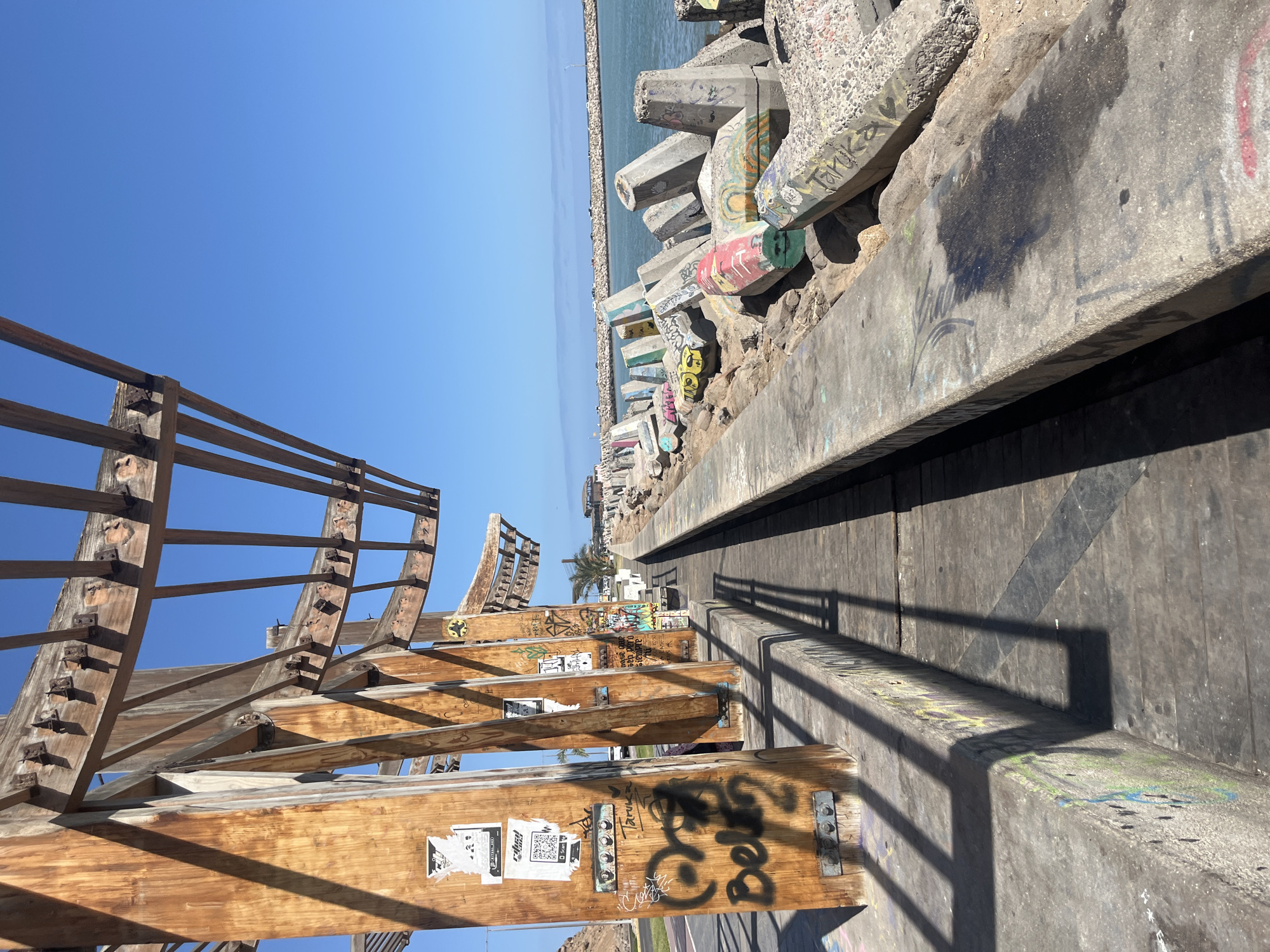 The image depicts a view from a wooden structure adorned with graffiti and stickers towards the Pacific Ocean in Arica, Chile. The structure, seemingly a partially constructed or decaying pier or viewing platform, is made of weathered wood and metal fasteners. The foreground features the structure's wooden beams and supports, heavily marked with graffiti and posters.
A concrete pathway or elevated walkway stretches into the distance. To the right of the walkway, there are large concrete tetrapods (coastal protection structures) stacked along the shoreline, also covered in graffiti. Beyond the tetrapods, the blue ocean extends to the horizon under a clear, cloudless sky. A faint line of land is visible in the distance. A palm tree is seen farther down the walkway. A street with railings is seen below on the left.