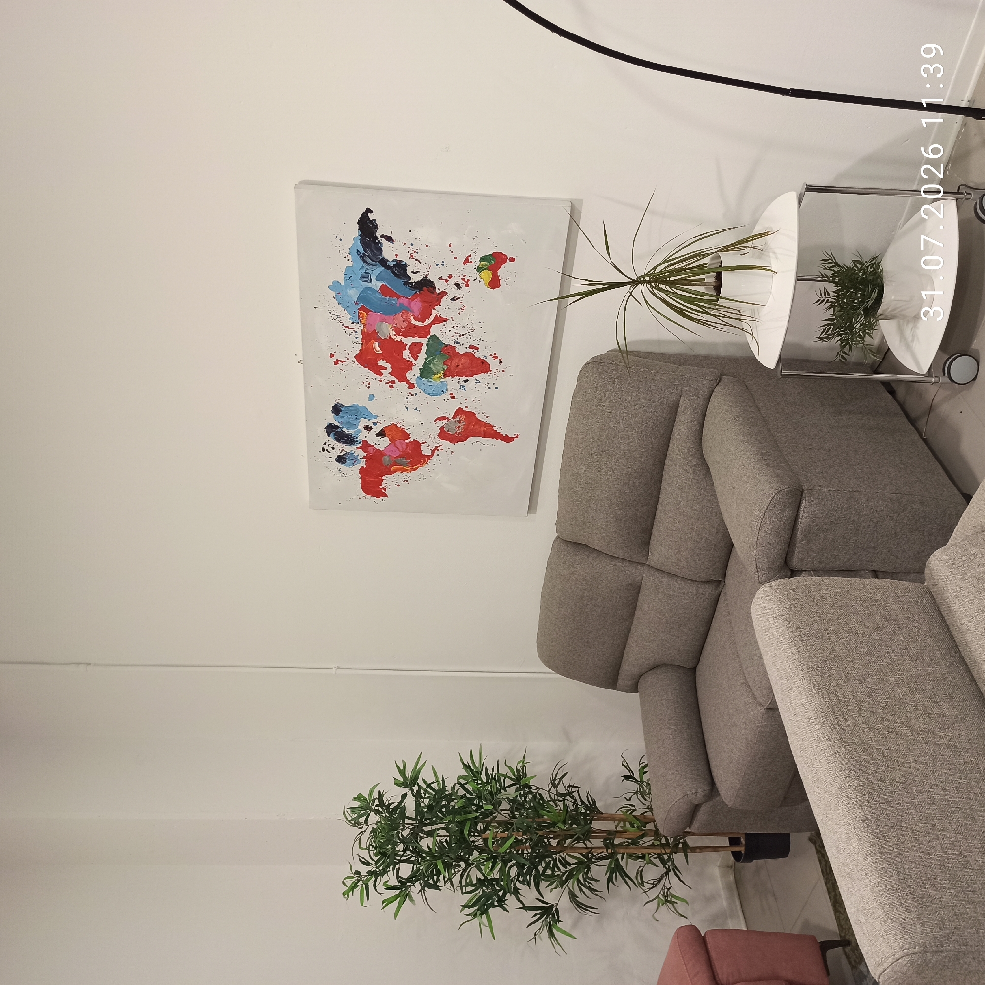 The image depicts an indoor scene, likely a living room or lounge area, with a minimalist aesthetic. The dominant elements are a gray sectional sofa, a tall artificial plant with bamboo-like stalks and green foliage, and a contemporary world map artwork on the wall. The artwork is abstract, featuring splashes of vibrant colors like red, blue, and yellow, against a white background, with the continents outlined. To the right of the sofa, there's a modern white side table with two tiers, each holding a potted green plant. A black arc floor lamp stands behind the side table. The walls are painted white, and the floor appears to be a light-colored tile or laminate. The overall impression is one of a clean, modern, and sparsely decorated space.
The timestamp "31.07.2026 11:39" is overlaid on the bottom right of the image.
The image does not provide any explicit information or visual cues that would allow for its specific location in the city of Modugno, Italy.