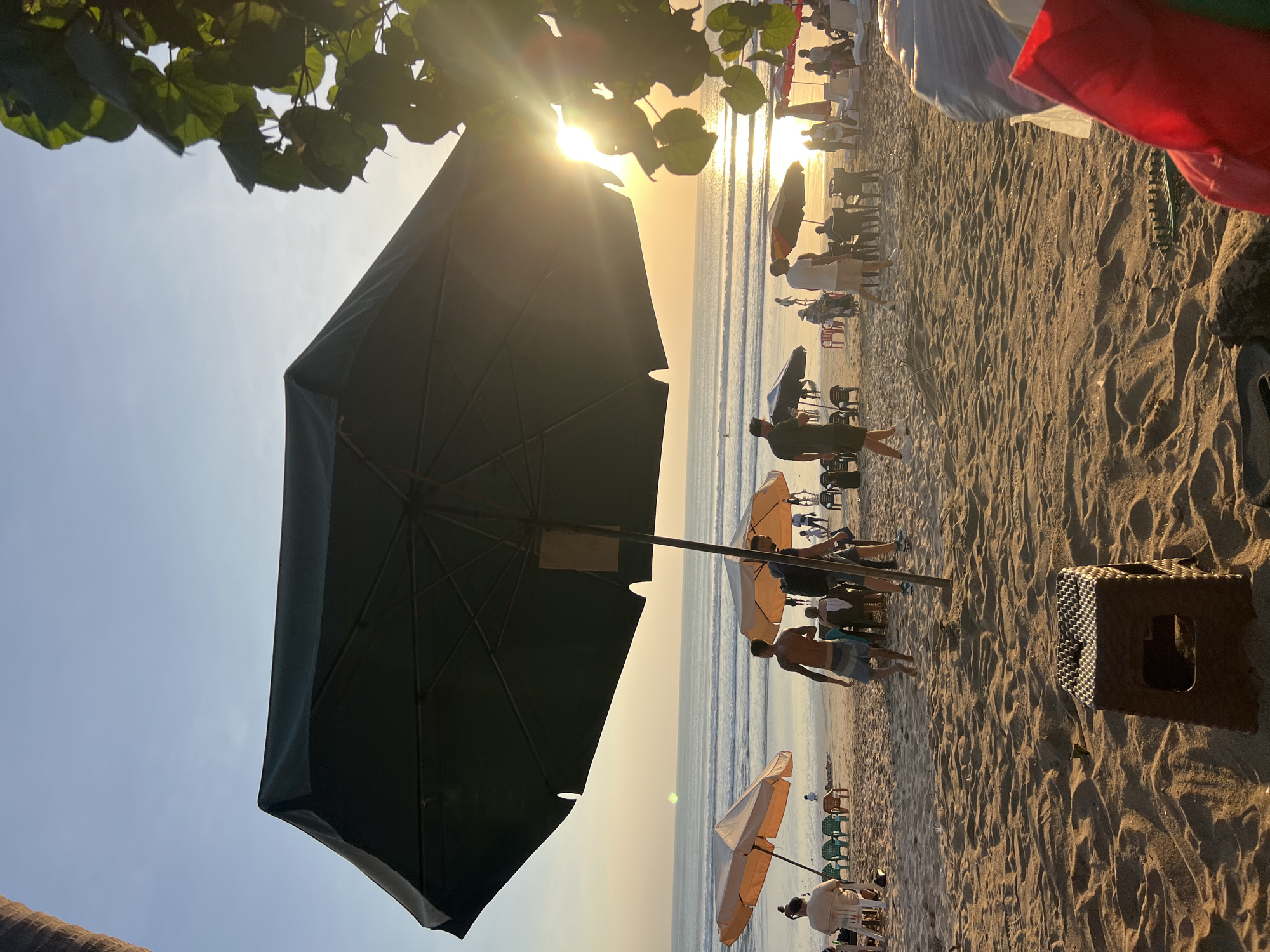 The image depicts a crowded beach scene in Kuta, Indonesia, during sunset. The sun is low in the sky, casting a warm, golden glow over the sand and water, with visible sun flares. The foreground is dominated by the sandy beach with the textured surface of the sand in focus, featuring a small, brown plastic stool and some discarded items on the right side.
In the mid-ground, several beach umbrellas are set up. A large, dark umbrella is prominent in the foreground, partially obscuring the view of the beach. To its left, two lighter-colored umbrellas stand over beach chairs. Further back, more umbrellas and beach furniture are visible, interspersed with groups of people.
The people on the beach are engaged in various activities. Some are walking along the shore, while others are seated under the umbrellas or on the sand. There are individuals in swim trunks and casual attire. In the background, the ocean displays gentle waves, and the horizon is a soft gradient of orange and blue sky. The presence of palm trees on the far right suggests the tropical setting of Kuta. The overall atmosphere is one of leisure and relaxation.