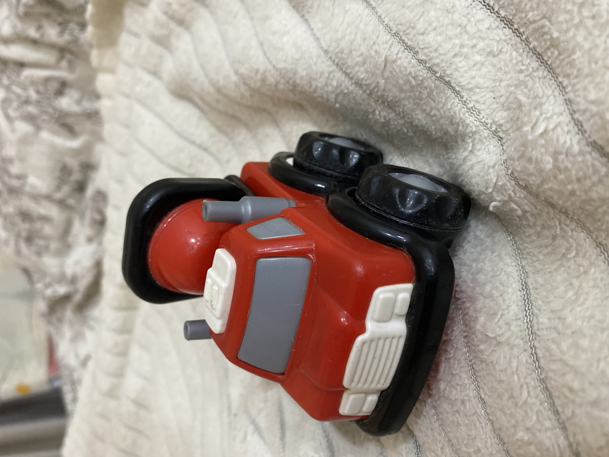 A red toy cement truck with black wheels and a black drum sits on a textured, light-colored blanket. The truck has a white grille, gray windows, and two gray exhaust pipes. The background is out of focus, showing more of the blanket and a hint of other objects. The image does not contain any information about the city of Balashikha, Russia, nor any people or actions.