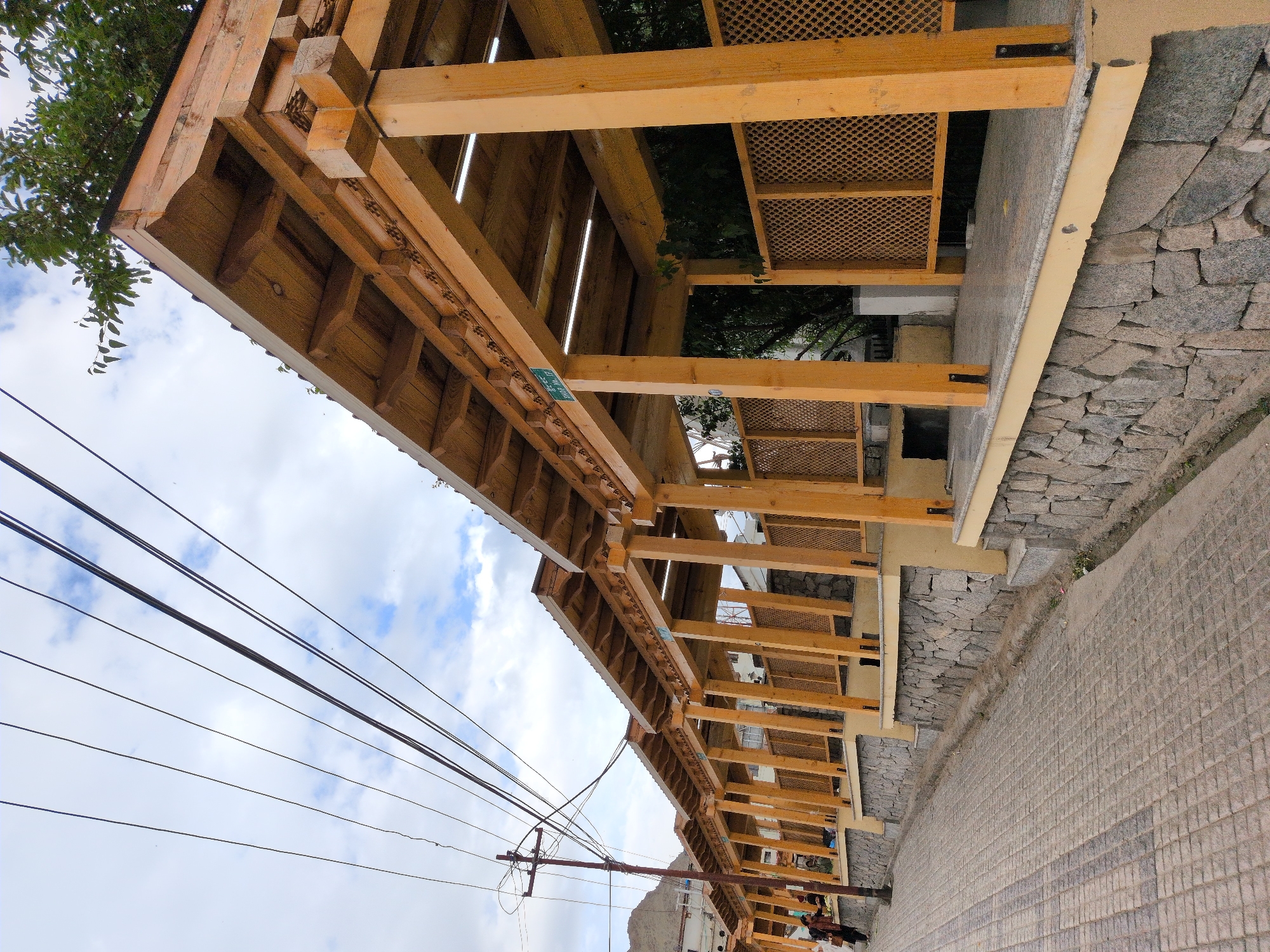 The image shows a covered walkway with wooden pillars and a sloped roof, situated in the city of Leh, India. The walkway is lined with a stone wall at its base and features decorative wooden latticework on the sides. A paved walkway with a cobblestone pattern runs alongside the covered structure. Overhead, a network of electrical wires crisscrosses the sky, which is partially obscured by clouds. In the background, buildings and a distant mountain are visible. A few individuals are present, one standing near the entrance of the walkway and another person further down. The overall scene depicts a functional and aesthetically designed public space in an outdoor urban environment.