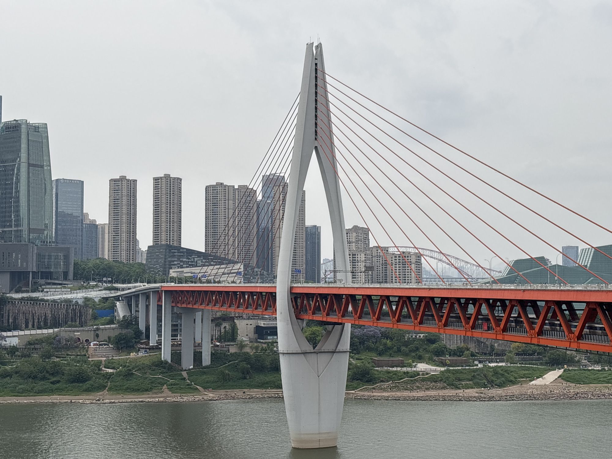 The image shows the Dongshuimen Bridge in Yuzhong District, Chongqing, China. The cable-stayed bridge has a distinctive white tower shaped like an inverted Y. The bridge deck is orange and has a lattice structure. Red cables extend from the top of the tower to support the deck.
In the background, there is a cityscape with several skyscrapers and other buildings. There is a river in the foreground. The riverbank is visible on both sides, with trees and some walkways. People are visible walking on the riverbank. Another bridge can be seen in the distance behind the Dongshuimen Bridge. The sky is overcast.