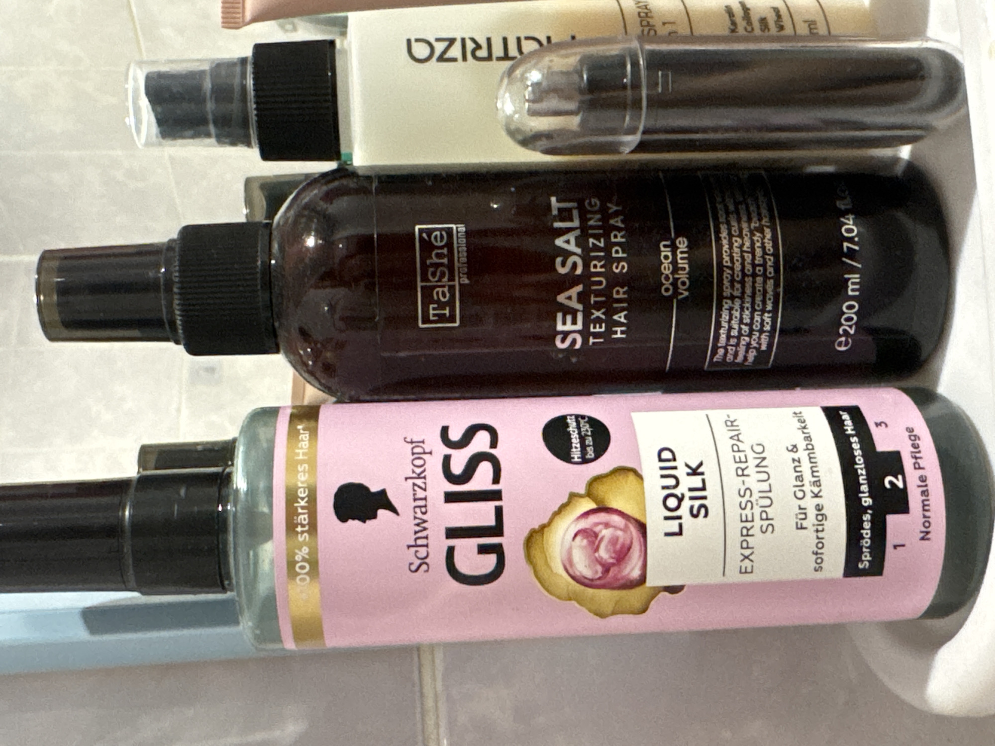 In the image, several bottles of hair products are displayed on a white shelf. The bottles are arranged in a line, with the one on the far left being the largest. The shelf appears to be in a bathroom or dressing area, with tiled walls in the background. The lighting is bright, suggesting it might be daytime. The bottles themselves are varied in color and design, with labels indicating different brands and product types. Some of the text on the labels is visible, including brand names like "Schwarzkopf Gliss" and "TaShé professional." One bottle has a pink label with a black silhouette of a person's head, and another has a dark brown bottle with "SEA SALT TEXTURIZING HAIR SPRAY" printed on it. There's also a lighter colored bottle with "RIZA" visible on the label, and a small pump bottle in front of it. The image is a close-up shot, focusing on the hair products. There are no people or animals visible in the image.