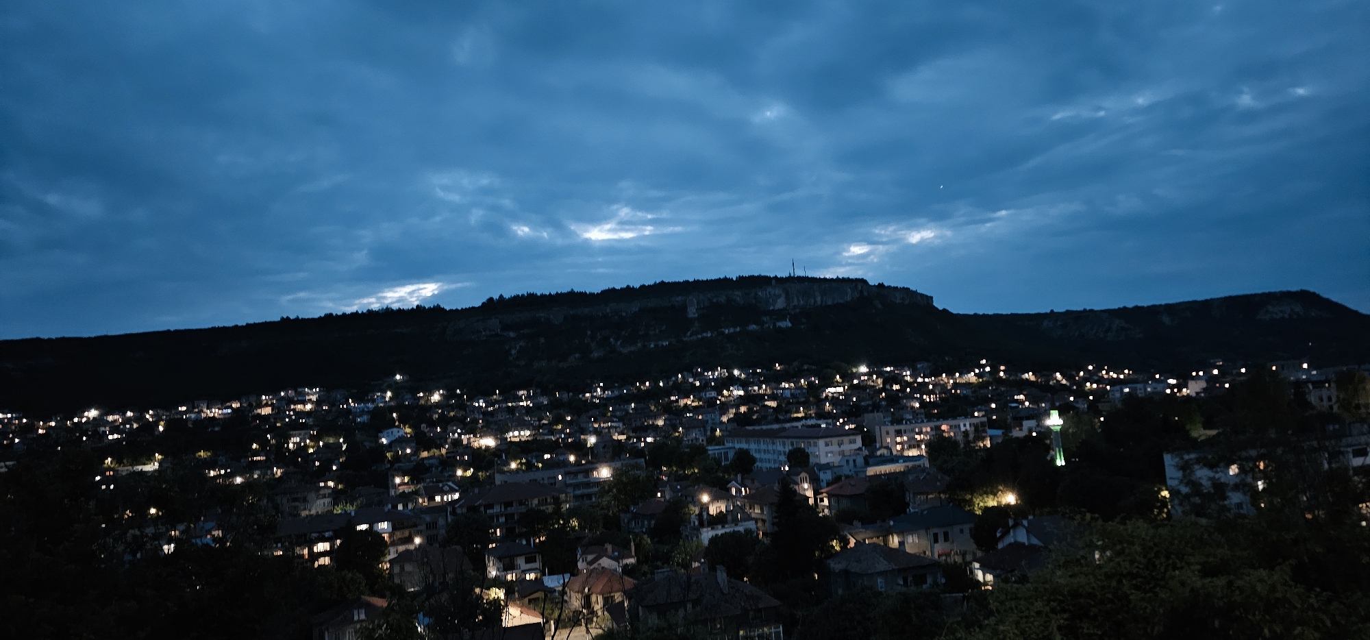 An elevated, wide-angle view captures a town nestled at the base of a significant hill or mountain under a twilight or nighttime sky. The sky is a deep, moody blue, with scattered clouds that show lighter, ethereal patches, suggesting the last glimmers of daylight or moonbeams.
The prominent hill in the background forms a dark, rugged silhouette, crowned with trees and featuring some visible rock formations. On its highest point, a slender structure, possibly an antenna or telecommunications tower, rises against the sky.
Below the hill, the sprawling town is illuminated by numerous artificial lights. These lights, emanating from houses and street lamps, appear as small, bright points of warmth against the surrounding darkness, signifying human presence and activity within the residential areas. The architecture suggests a European or Mediterranean style, with various rooflines and building shapes. One distinct feature in the town is a tall, slender structure resembling a mosque minaret, illuminated with a vibrant green light, visible towards the right side of the town.
The overall atmosphere is one of serene tranquility, characteristic of a quiet evening. There are no people visible, and no discernible text can be read in the image. The scene offers a peaceful panorama of a community glowing softly beneath the expanse of the dusk sky and the imposing natural landscape.