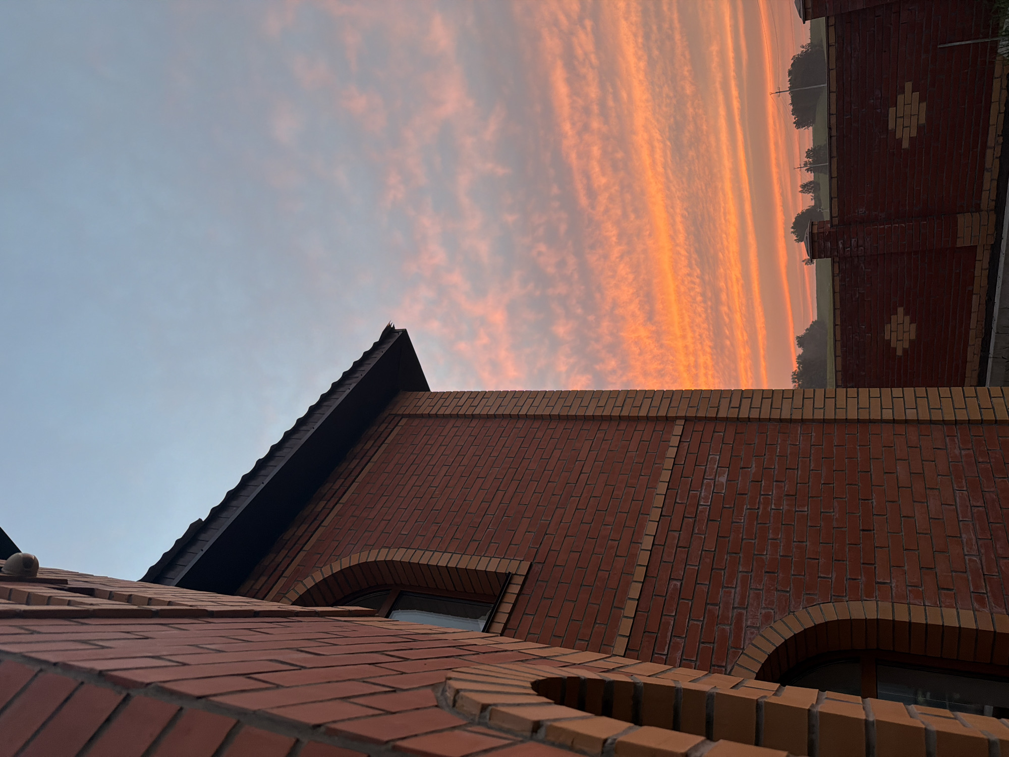 The image depicts the exterior of a red brick building with an arched window and a dark roof, seen from a low angle looking upwards. A section of a red brick fence with decorative diamond-shaped insets is visible in the lower right portion of the frame. Beyond the fence, there is a hazy landscape with trees and what appears to be a field. The sky is filled with striated clouds, predominantly in shades of orange and pink, suggesting either a sunrise or sunset. The scene is set in an outdoor environment, likely a residential or suburban area. The time of day is indicated by the colorful sky. There are no people or animals present in the image. The overall impression is of a quiet, picturesque moment during dawn or dusk. The location is specified as Krasnoyarsk, Russia.