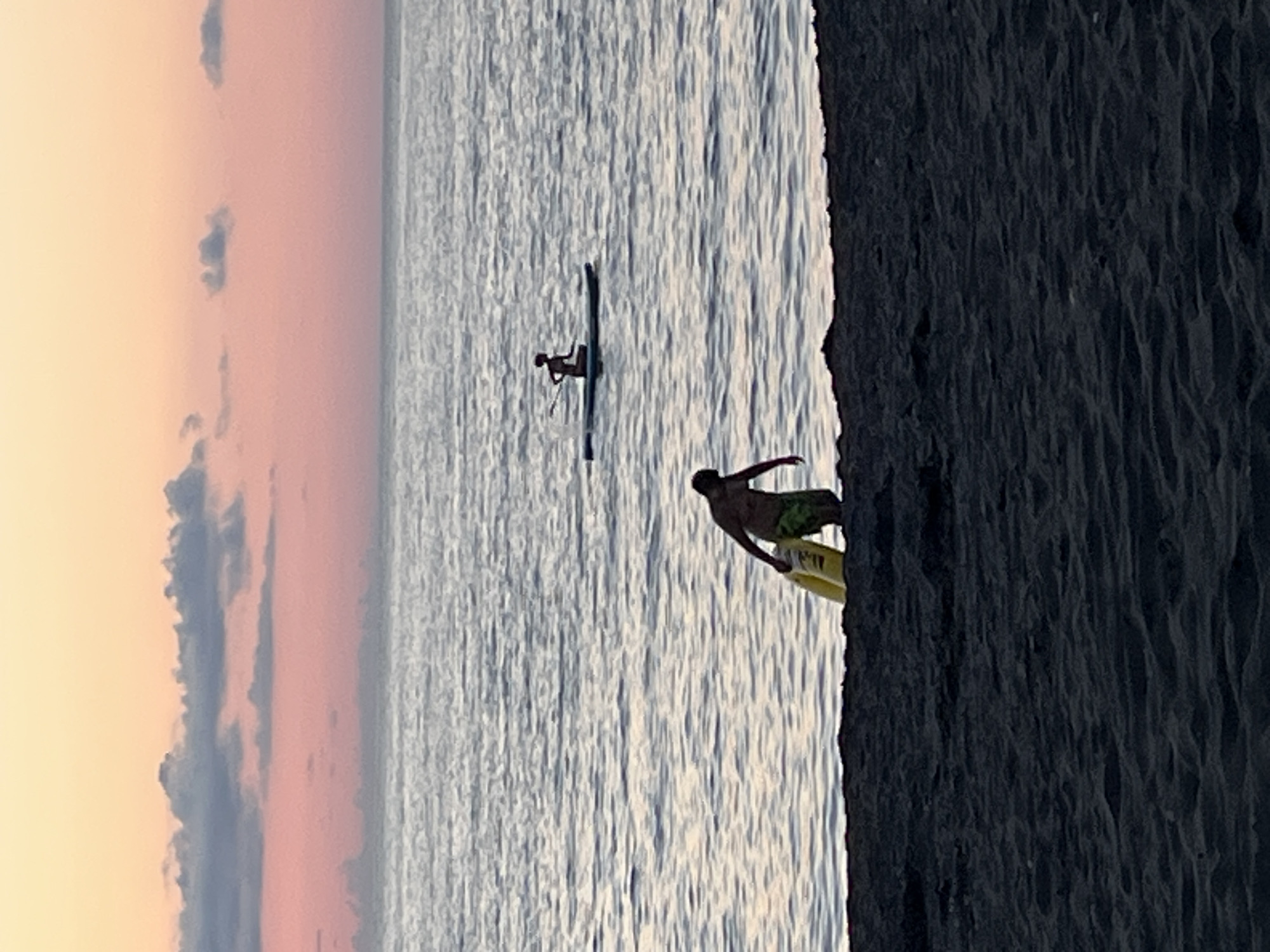 At dusk on the beach in Ureki, Georgia, a man is wading into the sea carrying a yellow flotation device. Further out on the water, a person is seated on a paddleboard, holding a paddle. The sky is a gradient of pink and orange, with a few dark clouds, and the sea is calm with gentle ripples. The foreground shows a dark sand beach.