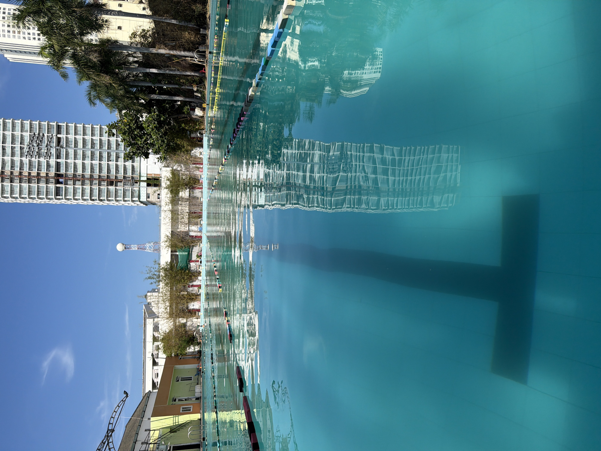 This media file captures a vibrant outdoor scene featuring a large swimming pool under a clear blue sky, set against a backdrop of urban architecture and palm trees. The image is oriented vertically, showing the pool on the right and the surrounding landscape on the left.
The foreground is dominated by the swimming pool, filled with clear, turquoise-blue water. The water's surface acts as a mirror, reflecting the tall buildings and sky. Colorful lane lines with floats extend across the pool. Below the water's surface, a large, dark, cross-shaped marking is visible on the bottom of the pool, indicating its depth or a specific area.
To the left of the pool, under a bright, cloudless blue sky, stand several tall, modern high-rise buildings. One prominent building is white with a grid-like pattern of numerous windows. Interspersed among the buildings are several tall palm trees, adding a tropical touch to the urban setting. Further in the background, there are lower, more modest buildings, including one painted in shades of yellow and green, and a distinctive white dome structure atop a tower, possibly a radar or telecommunications antenna. A crane-like structure is also visible in the distance.
The scene is bathed in bright daylight, suggesting a sunny day. No people or specific activities are visible, presenting a serene view of the pool area. The combination of modern high-rises, palm trees, and the clear blue pool water aligns with the tropical urban environment of Nha Trang, Vietnam. There is no discernible text visible in the image.