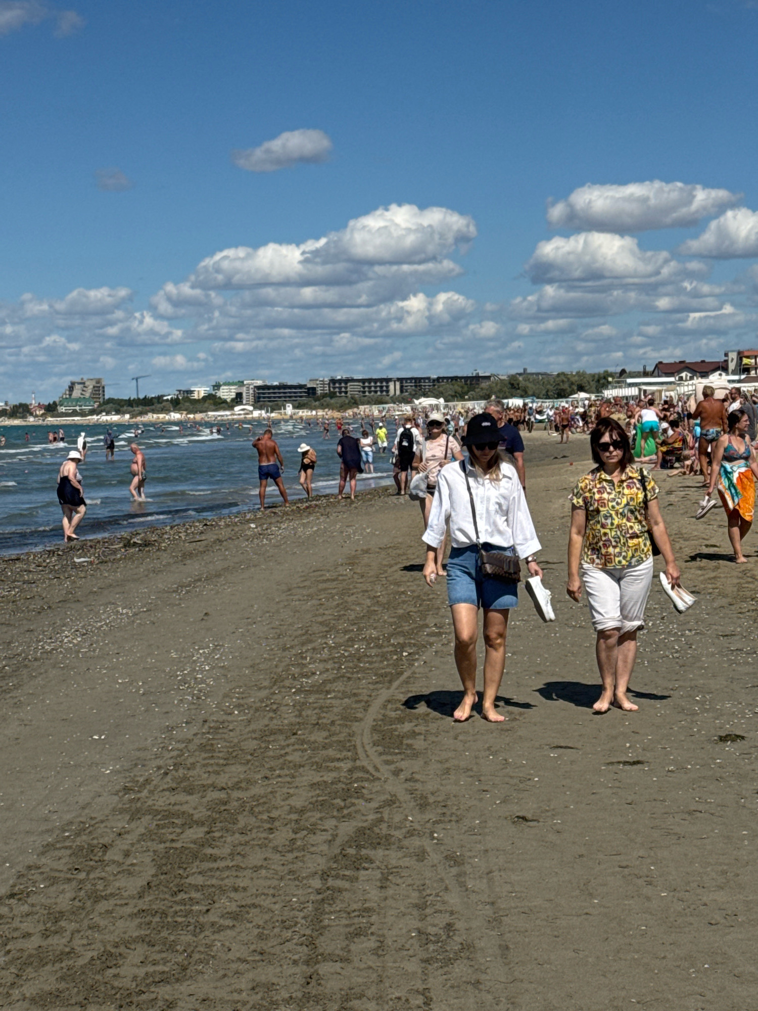 The image depicts a beach scene in Anapa, Russia, under a partly cloudy sky. The foreground shows a wet sand beach with a line of shells and debris. Several people are visible walking on the beach and in the shallow water. In the middle ground, two women walk towards the viewer, carrying their shoes. One is wearing a white shirt and denim shorts, the other a colorful patterned shirt and white shorts. Behind them, a crowd of beachgoers are seen walking, relaxing on the sand, and in the water. The background reveals the coastline with numerous buildings, including hotels and other structures, suggesting a developed resort area. The Black Sea is visible with gentle waves.