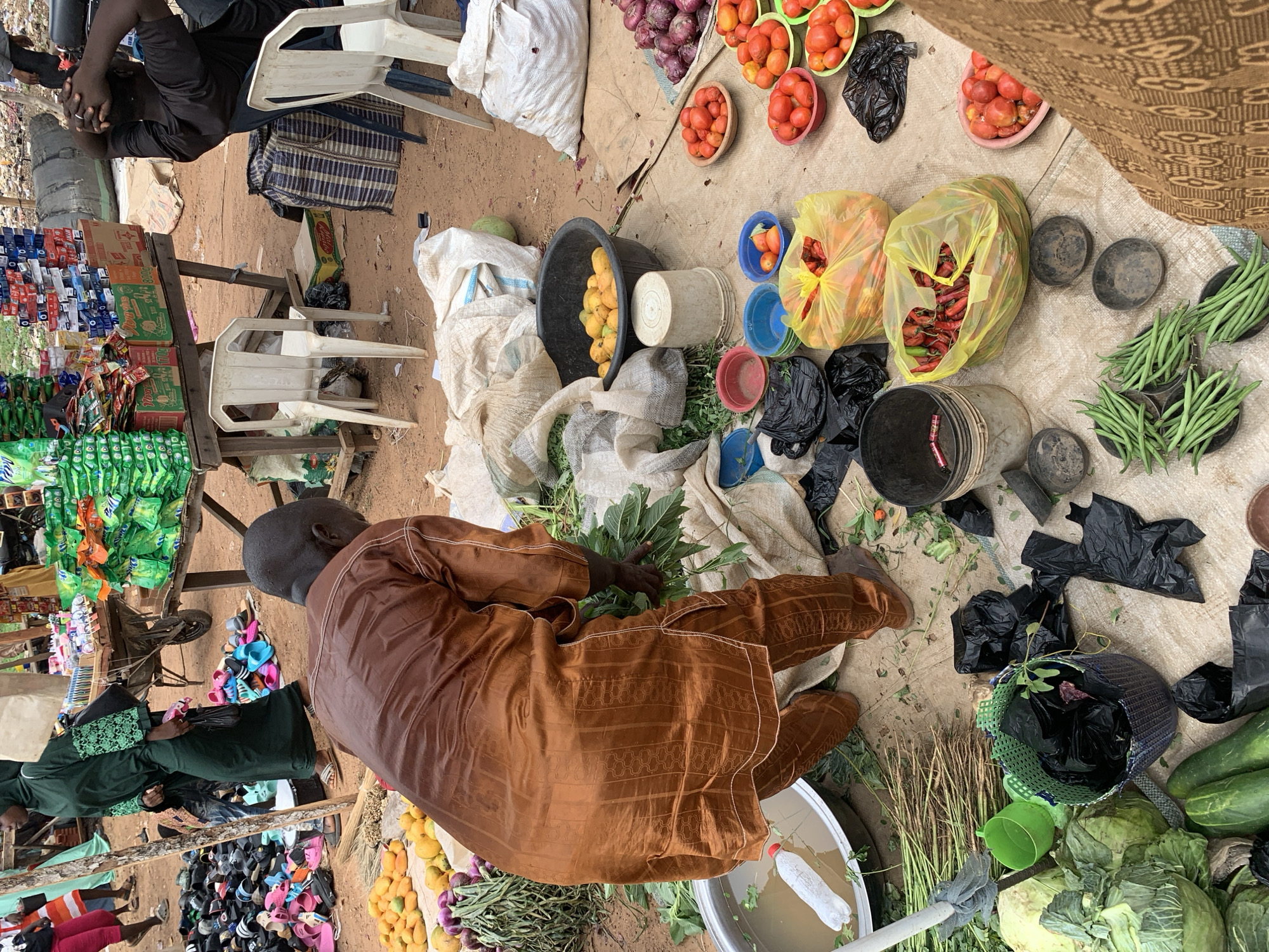 This overhead view captures a vibrant outdoor market scene in Keffi, Nigeria, showcasing an array of fresh produce and various goods. The ground is dusty earth, with items laid out on large light-colored fabric sheets or on makeshift wooden stalls.
In the foreground, a man wearing a brown traditional long-sleeved garment is seated on the ground, facing away from the camera. He is engaged with fresh leafy greens spread before him, seemingly sorting or preparing them. Next to him, a white plastic water bottle is partially submerged in a large basin of water.
The market displays a rich variety of produce: numerous red and orange tomatoes are piled in colorful plastic bowls and black plastic bags. Piles of red onions and yellow fruits (possibly mangoes) are visible in a black basin. Green beans are neatly arranged in metal bowls, while cucumbers and large heads of cabbage are also displayed. Bright red chili peppers are packed into transparent yellow plastic bags.
To the left, a wooden stall holds various packaged goods, including green sachets with the brand "Bounty" and boxes labeled "Always," "Safecare," "Golden Penny," and "MAGGI." A woman in a green abaya-like dress stands near this stall, observing the merchandise. In the background, another man in a black t-shirt sits on a white plastic chair, appearing to rest with his head down. Colorful sandals and slippers are laid out on the ground near another stall. Scattered throughout the market are black plastic bags, burlap sacks, and assorted plastic basins and buckets.
The scene is brightly lit, indicating daytime under clear weather conditions. The overall atmosphere is that of a calm, bustling local market. No strong emotions are discernible from the individuals.