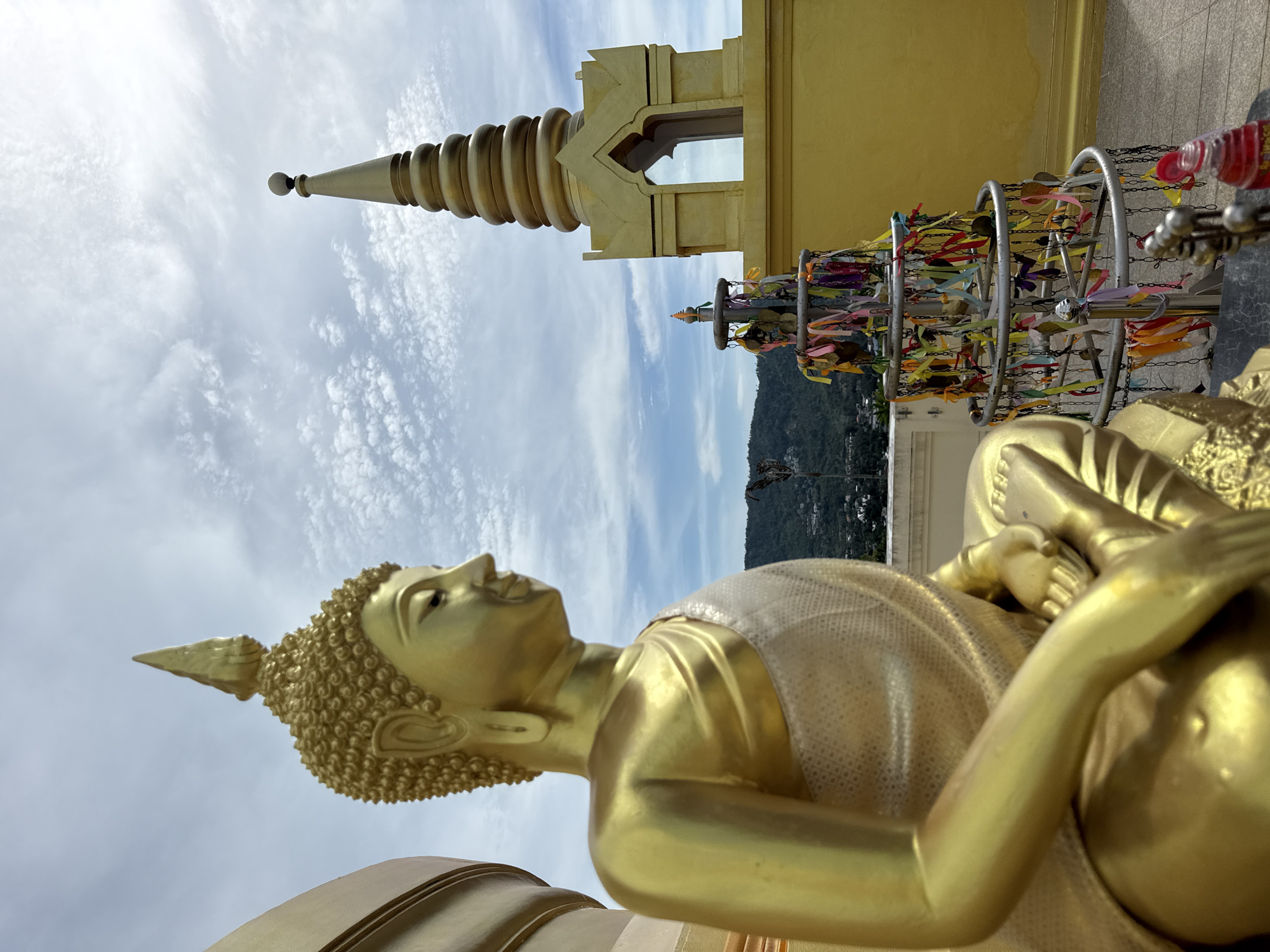 A golden Buddha statue sits in meditation outdoors, in front of a golden stupa and against a cloudy sky. The statue is positioned in the foreground, with its left side and profile facing the camera. It wears a flowing robe draped over its left shoulder, adorned with an intricate pattern. Behind the Buddha, a tiered, golden stupa rises, topped with a spire. In the midground, a circular metal structure is decorated with numerous colorful ribbons and small charms. The background shows a lush green hillside dotted with buildings, suggesting the location is within or near a populated area. The overall scene is serene and spiritual.