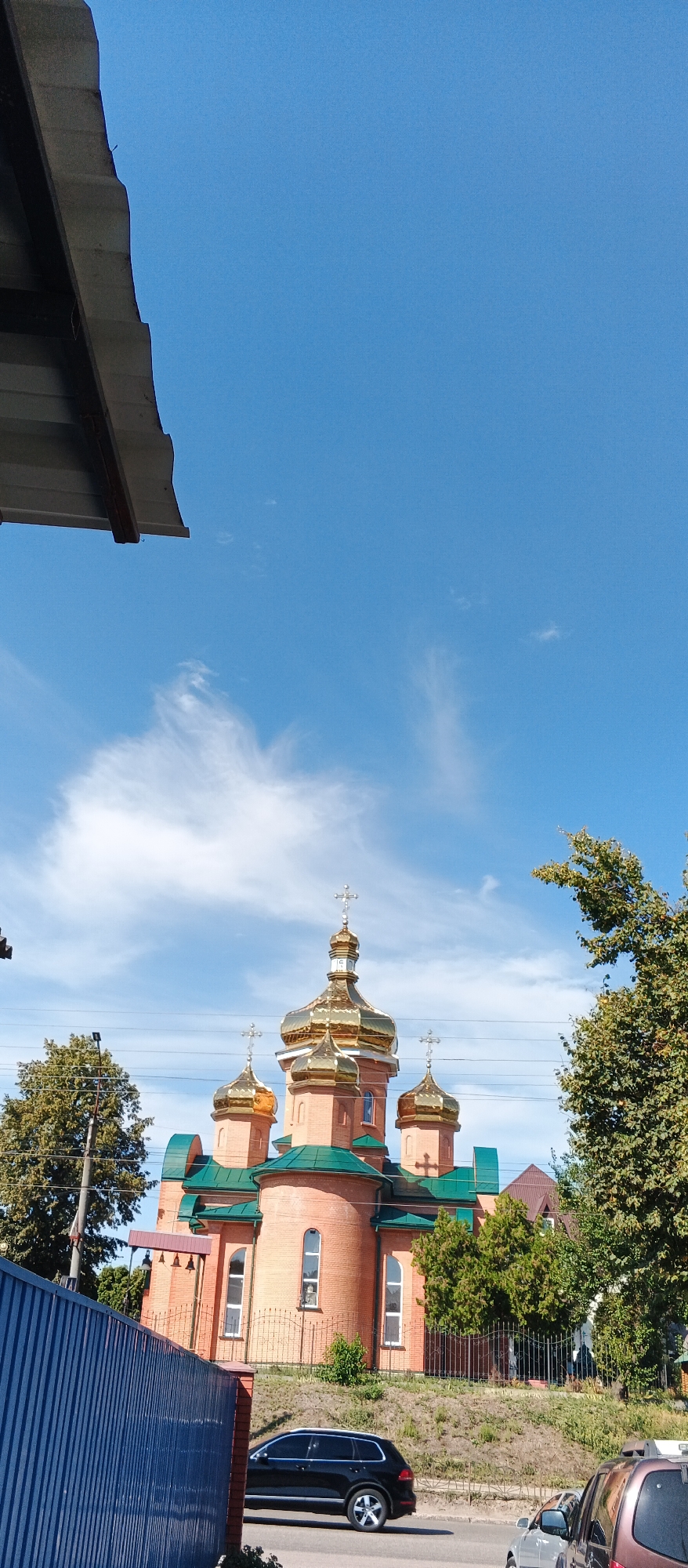 A three-quarter-profile, eye-level, outdoor, daytime shot shows the Church of the Intercession in the city of Smila, Ukraine, viewed from a roadside across a blue metal fence. The church is constructed of red brick with green roofing and multiple golden domes topped with crosses. A black SUV is parked to the right of the church, and a portion of another vehicle is visible on the far right. The sky is a clear blue with wispy white clouds. Trees line the background of the church, and a grey overhang is visible in the upper left corner of the frame.