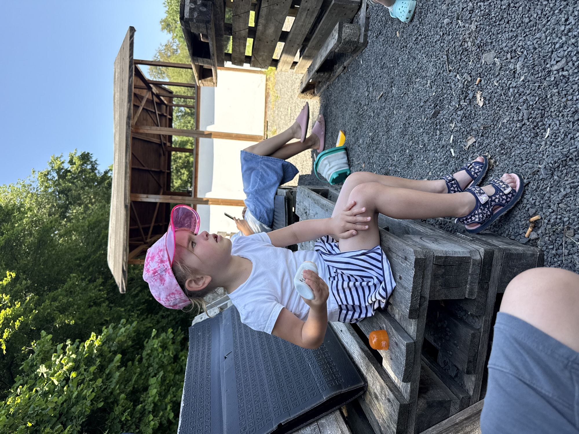 An outdoor scene in Beltersheim, Germany, features a young girl sitting on a wooden pallet bench. She is wearing a white t-shirt, a pink patterned baseball cap with a clear brim, and a black and white striped skirt. The girl is holding a white cylindrical object in her right hand. To her left, on the pallet, is an orange, round object with a label. Another person, out of focus, is visible sitting behind her on a similar pallet bench, wearing a blue skirt or dress and pink slippers. The environment is characterized by a gravel ground, other wooden pallets stacked nearby, and trees and a simple wooden shelter in the background under a clear blue sky.