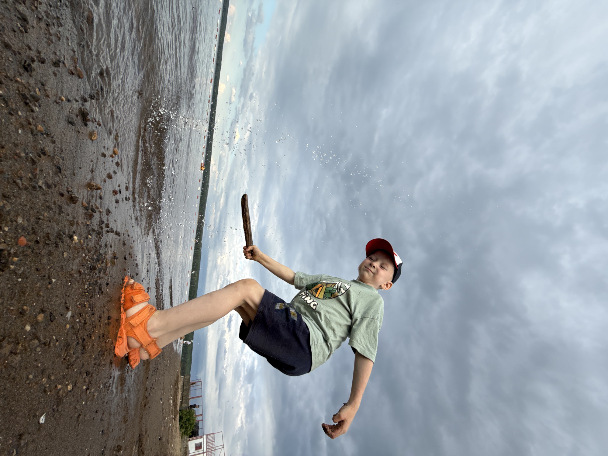 A young boy in a green t-shirt, dark shorts, and orange sandals is depicted mid-air, as if jumping or being thrown. He holds a stick in his right hand. His face is tilted upwards, eyes closed, and a smile suggests he is enjoying himself. He wears a red and black baseball cap. The background consists of a cloudy sky. The foreground shows a beach with wet sand and small pebbles, with water lapping at the shore. Splashes of water are visible. In the distance, on the left side of the image, a small structure and some trees are visible along the shoreline. The overall impression is one of playful activity on a beach. The time of day appears to be overcast, likely late afternoon or evening. The location is identified as Chaikovsky, Russia. The t-shirt has a graphic with the word "PING" visible.