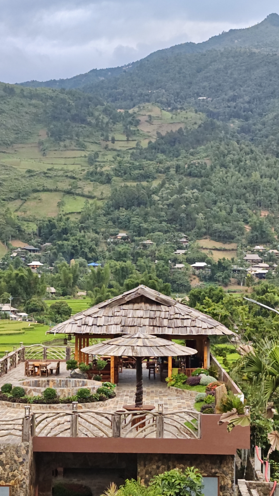A vertical, full-shot image captures a building with a wooden-shingled roof, likely a gazebo or pavilion, overlooking a landscape in Tú Lệ, Vietnam.
The building is elevated on what appears to be a stone platform, adorned with an intricately designed wooden railing. The roof is the building’s most distinctive feature, crafted from numerous overlapping wooden shingles. A second, smaller roofed structure is present within the larger one, supported by a wooden post. Tables and chairs are visible within the structure.
The building overlooks a vast landscape of tiered rice paddies and a densely populated settlement. Houses are scattered among lush greenery. Behind the settlement, rolling hills covered in verdant vegetation dominate the background. The sky above is overcast, suggesting a cloudy day.