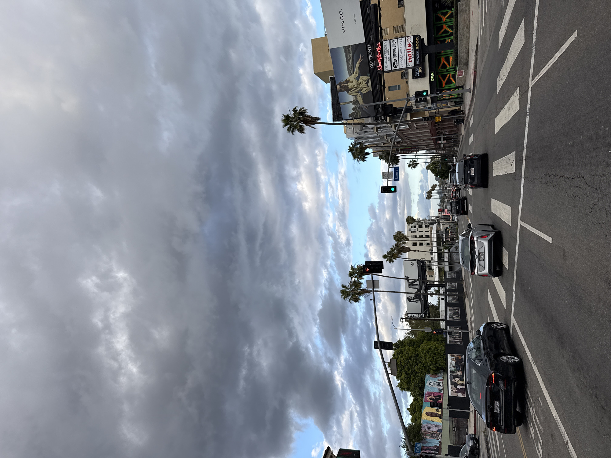 The image depicts a street scene in Los Angeles, California, USA, with a view along a north-south oriented avenue under a heavily clouded sky. Palm trees line the street, characteristic of the region. A street crossing is visible in the foreground, with several cars moving along the road. The street sign reads "Sunset Laurel". The buildings lining the street display various businesses and billboards, including signs for "VROMAGE CUISINE", "SMOKE SHOP", "VIVOLI CAFÉ", "BUBURGER 3000", "NAILS one", "SUPERSTAR" and "VINCE". One sign contains the text "OUTFRONT/ Sunset Laurel". The traffic lights indicate that the light is green for traffic moving in the photographed direction. Murals are painted on some of the building walls. There are no people prominently featured in the scene.