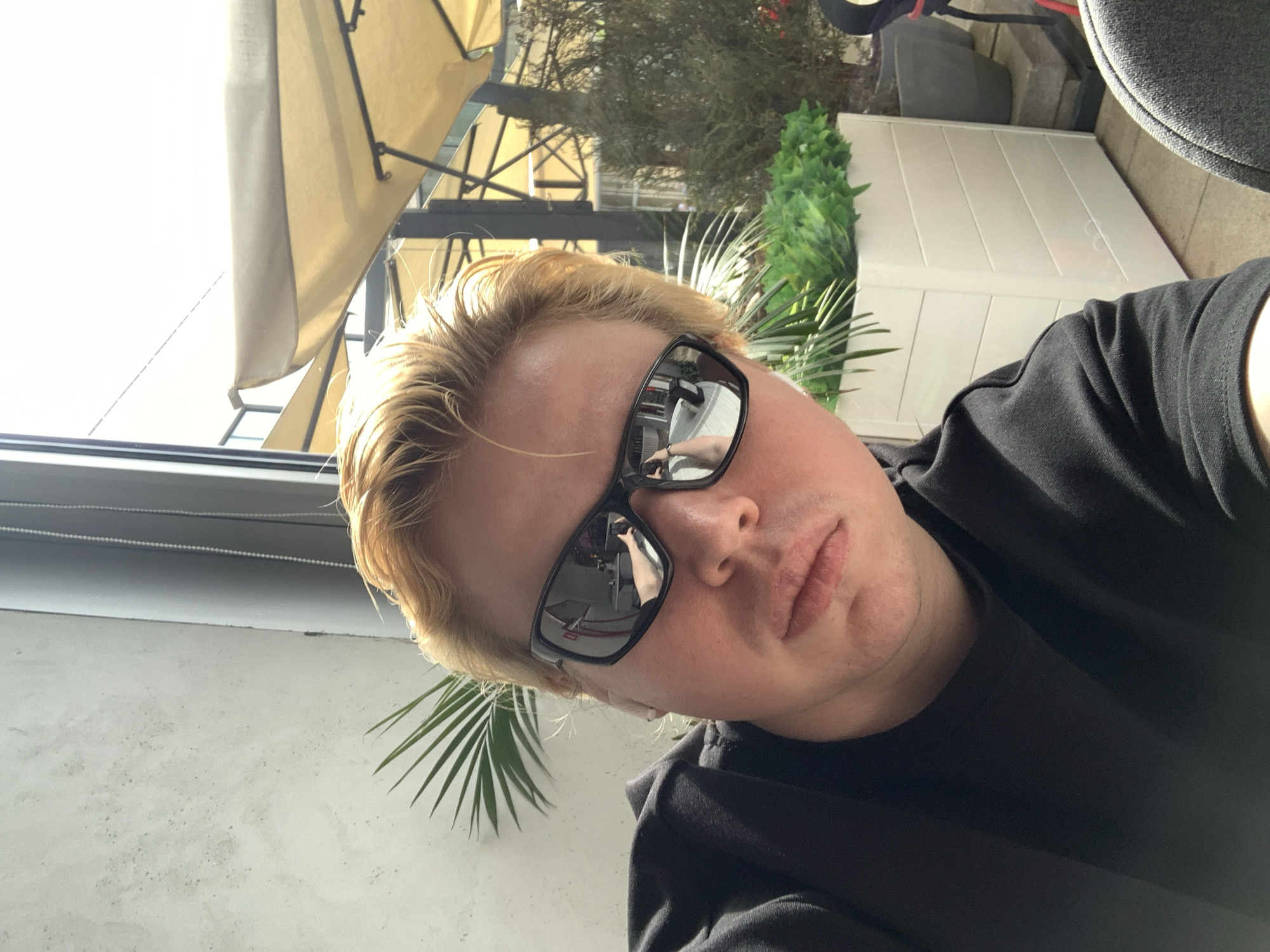 A Caucasian male with short, blonde hair and wearing black sunglasses and a black t-shirt is the primary subject of the image. The sunglasses reflect the act of taking the photo. The location appears to be an outdoor seating area. An open window is to the left, while a building's outer wall is to the right. There is an umbrella awning above. Green plants are visible in the background. The exact location in Almaty, Kazakhstan, cannot be definitively determined.