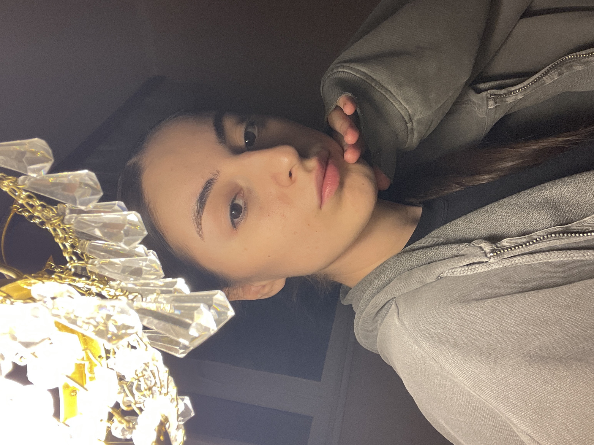A close-up indoor photograph depicts a young female, likely of Eastern European or Middle Eastern descent, looking directly towards the camera. She possesses dark, pulled-back hair, dark eyes, a prominent nose, and full lips, displaying a neutral to slightly pensive expression. Her right hand, with fingers curled, rests beneath her chin. She is dressed in a light gray or olive-green zippered hooded sweatshirt over a dark undergarment. In the upper left quadrant, a brightly illuminated chandelier with a gold-toned frame and multiple clear, faceted crystal pendants serves as a primary light source, creating significant glare and lens flare. The immediate background is dark and indistinct, offering no specific environmental details. No identifiable architectural features, landmarks, or unique objects are present to determine a specific location within Moscow, Russia.