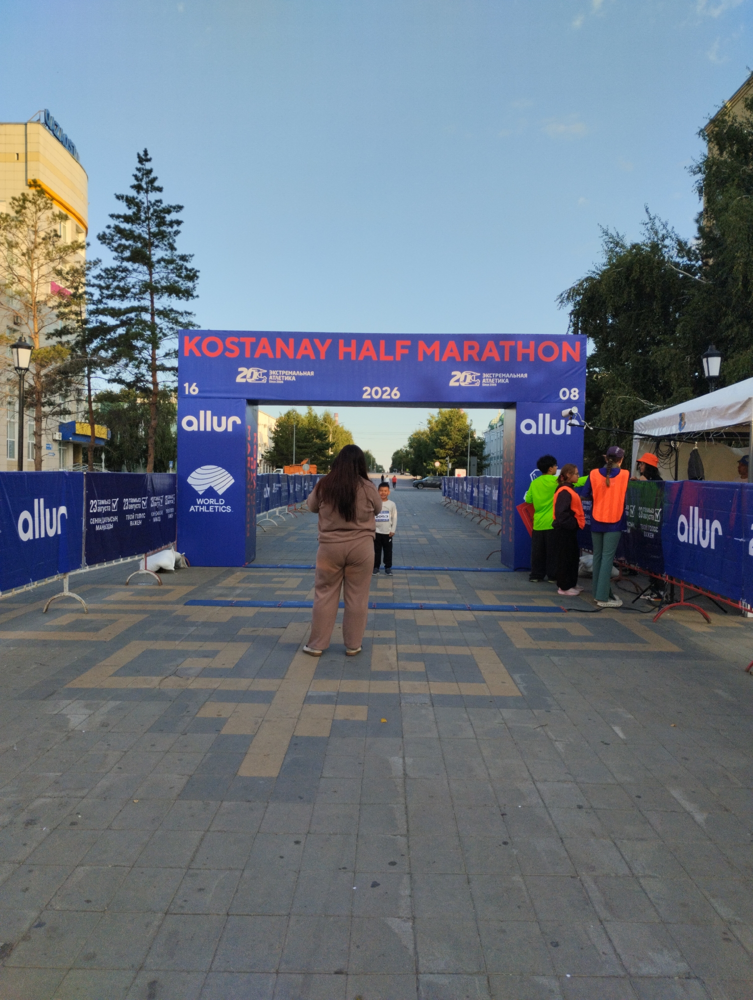 The image shows a street scene in Kostanay, Kazakhstan, with a large blue archway labeled "KOSTANAY HALF MARATHON". The archway is decorated with logos of "Allur" and "World Athletics". Several people are gathered around the archway, some wearing race bibs and athletic wear. There are also banners and barriers set up along the street, indicating a race event. The environment appears to be an urban setting, with buildings and trees visible in the background. The sky is clear and blue, suggesting good weather for the event.