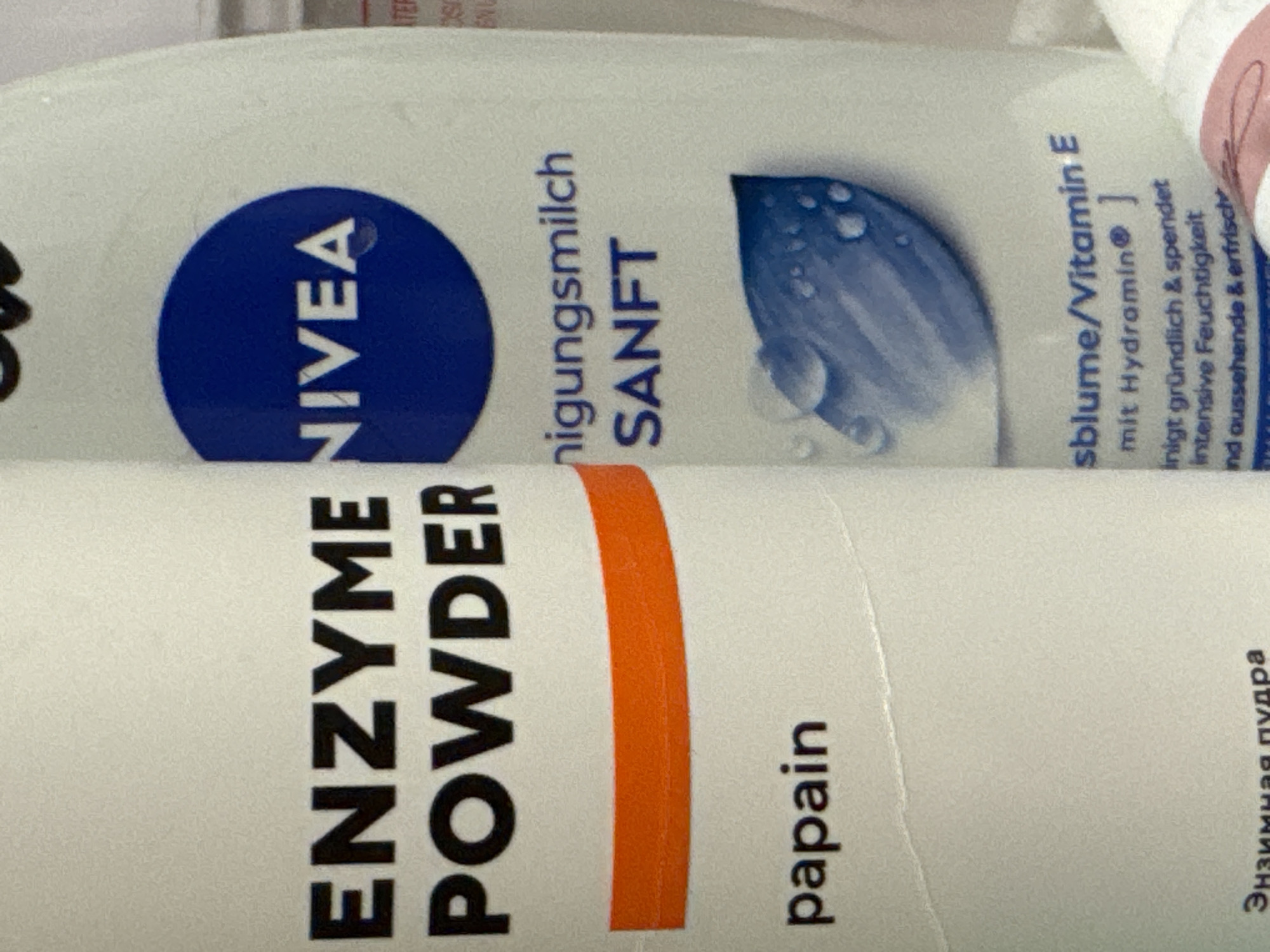 This is a close-up, overhead shot of two cosmetic product bottles, likely in a bathroom or on a shelf. The bottles are white with prominent branding. The upper bottle features the Nivea logo in blue, along with text in German that reads "Reinigungsmilch SANFT" and "sblume/Vitamin E mit Hydramin". Further German text describes the product's benefits. The lower bottle has bold black text in English, "ENZYME POWDER," and "papain." There is also Russian text visible on the lower bottle that translates to "Enzyme powder." The lighting suggests an indoor setting.