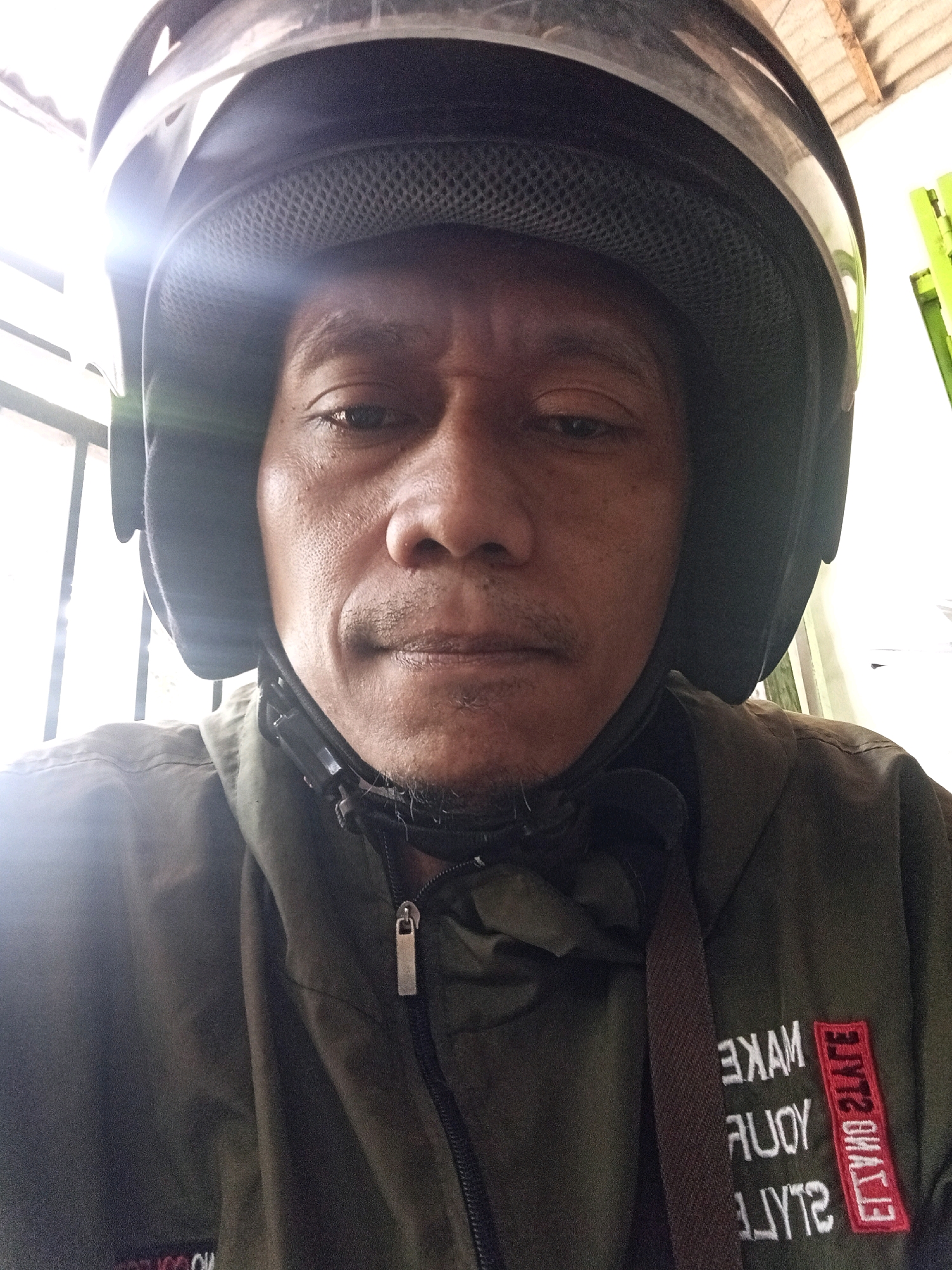 The image is a close-up, selfie-style photograph of an Indonesian man wearing a black helmet and a dark green jacket. The man is facing the camera, with his head slightly tilted. He has a light mustache and a goatee. The jacket has white text embroidered on the left side, which appears to be a reversed English phrase. The background is slightly blurred, showing what appears to be a building with wooden beams and possibly metal bars or a fence. The overall lighting suggests an outdoor or semi-outdoor setting, possibly during the day. The provided hints indicate the location is Wates, Indonesia.