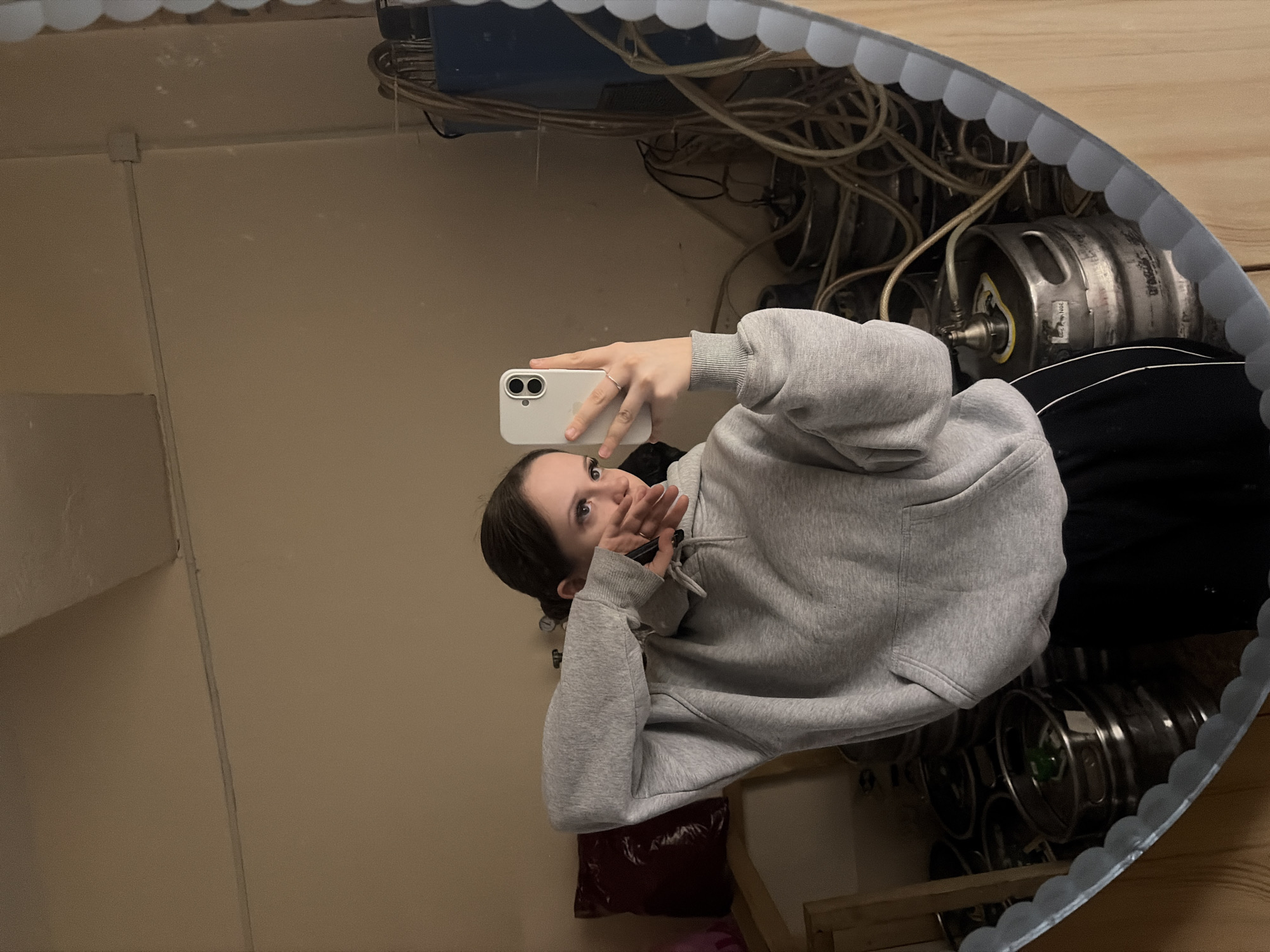 A young person is captured in a mirror selfie, reflecting an indoor setting. The individual, appearing to be female, is wearing a grey hooded sweatshirt and black trousers with a white stripe. They hold a white iPhone with a dual-camera setup, looking directly at the phone's camera lens. One hand holds the phone, while the other is raised to cover part of their mouth, conveying a playful or slightly shy expression with wide eyes.
The background, visible in the mirror's reflection, reveals what appears to be a storage or utility area. Numerous metal kegs, likely for beverages such as beer, are stacked and arranged. One keg clearly shows the text "30L", indicating its capacity. Tangled bundles of cables and hoses are visible, along with industrial-style connections leading to the kegs. The walls are a plain, light beige color, with some conduit running along them. A blue object is also partially visible among the cables. The mirror itself has a distinctive scalloped or wavy decorative edge. The setting suggests a back room of a commercial establishment like a pub, bar, or brewery in Semyonov, Russia.