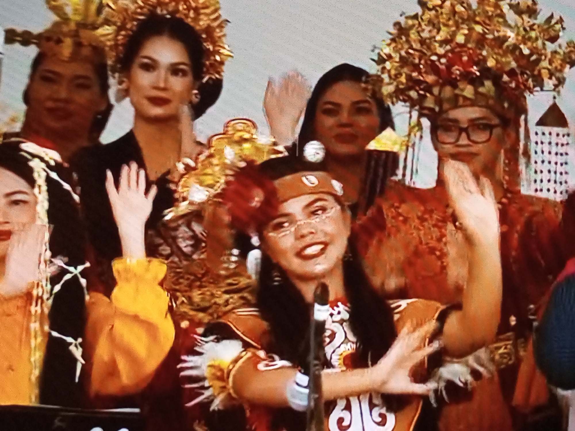The image shows a group of women in elaborate, traditional attire, likely participating in a cultural event. The woman in the foreground, center, is wearing a golden headdress adorned with red and gold floral elements. She has dark hair, noticeable makeup, and is wearing glasses. She is dressed in a red garment with intricate white and gold patterns. Her arms are raised, and her hands are in a gestural pose. To her left, another woman in a black top and a golden headdress is partially visible. Behind them, other women can be seen, also in traditional dress and headdresses. The setting appears to be indoors, possibly a stage or a formal gathering space, with decorative elements that suggest a festive atmosphere. The overall impression is of a vibrant cultural celebration. There is no information available to specify the exact location in Banyudono, Indonesia.