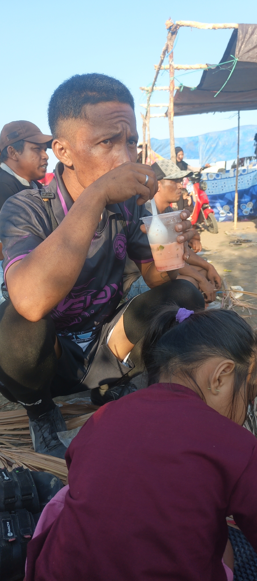 In the city of Lori, Indonesia, under a clear blue sky, a man is captured in a medium shot as he drinks from a plastic cup containing a pink beverage with visible chunks of fruit. He is crouched, wearing a dark grey and purple athletic shirt, black compression pants, and grey sneakers. A ring is visible on his right hand. In the foreground, the back of a young girl wearing a maroon shirt is visible. Behind the main subject, another man in a brown cap and a woman in a headscarf are slightly out of focus. A red scooter and a blue tarp structure are visible in the background, suggesting an outdoor setting, possibly a market or gathering area. The ground is covered with dry palm leaves.