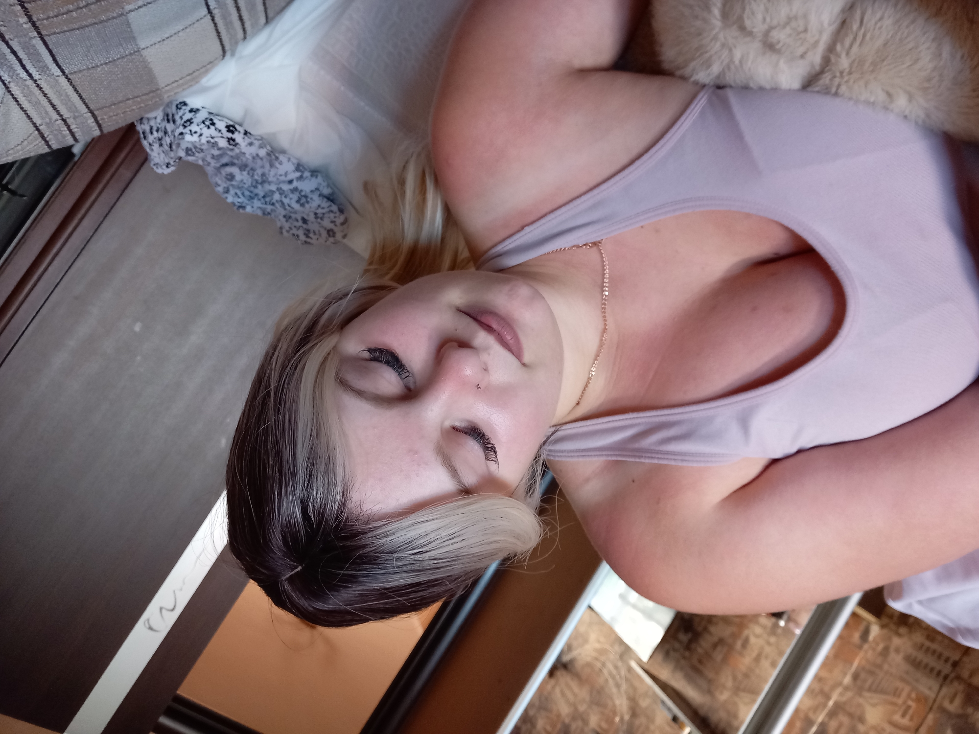 A young woman with blonde and dark hair, and fake eyelashes, is lying down indoors, looking down with her eyes closed. She is wearing a light purple tank top and a delicate necklace. The background consists of furniture and a patterned fabric. The lighting suggests it is daytime.