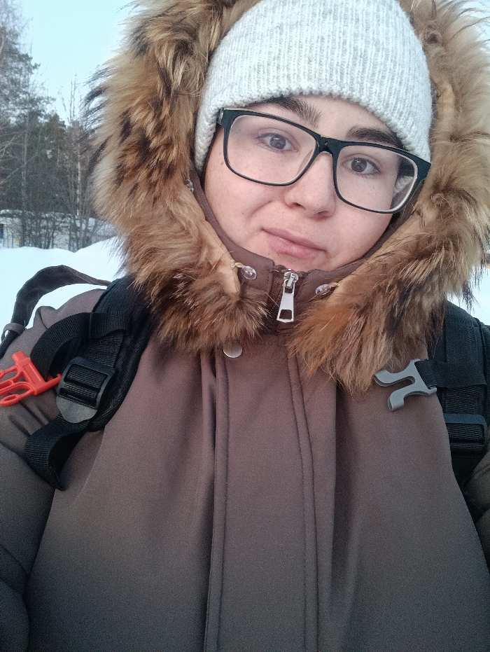 A close-up, eye-level portrait shot captures an individual outdoors in a snow-covered area identified as Noviy, Russia. The subject, with dark hair, wears a light grey knitted beanie and black-framed eyeglasses. A brown winter parka with a substantial brown fur-lined hood is worn, partially zipped to reveal a metallic zipper pull. Black backpack straps are visible over both shoulders; the left strap features a red plastic clip, and the right strap has a dark grey plastic buckle. The individual maintains direct eye contact with the camera, exhibiting a neutral expression. The background consists of snow-covered ground and a sparse arrangement of bare trees under a bright, possibly overcast, sky.