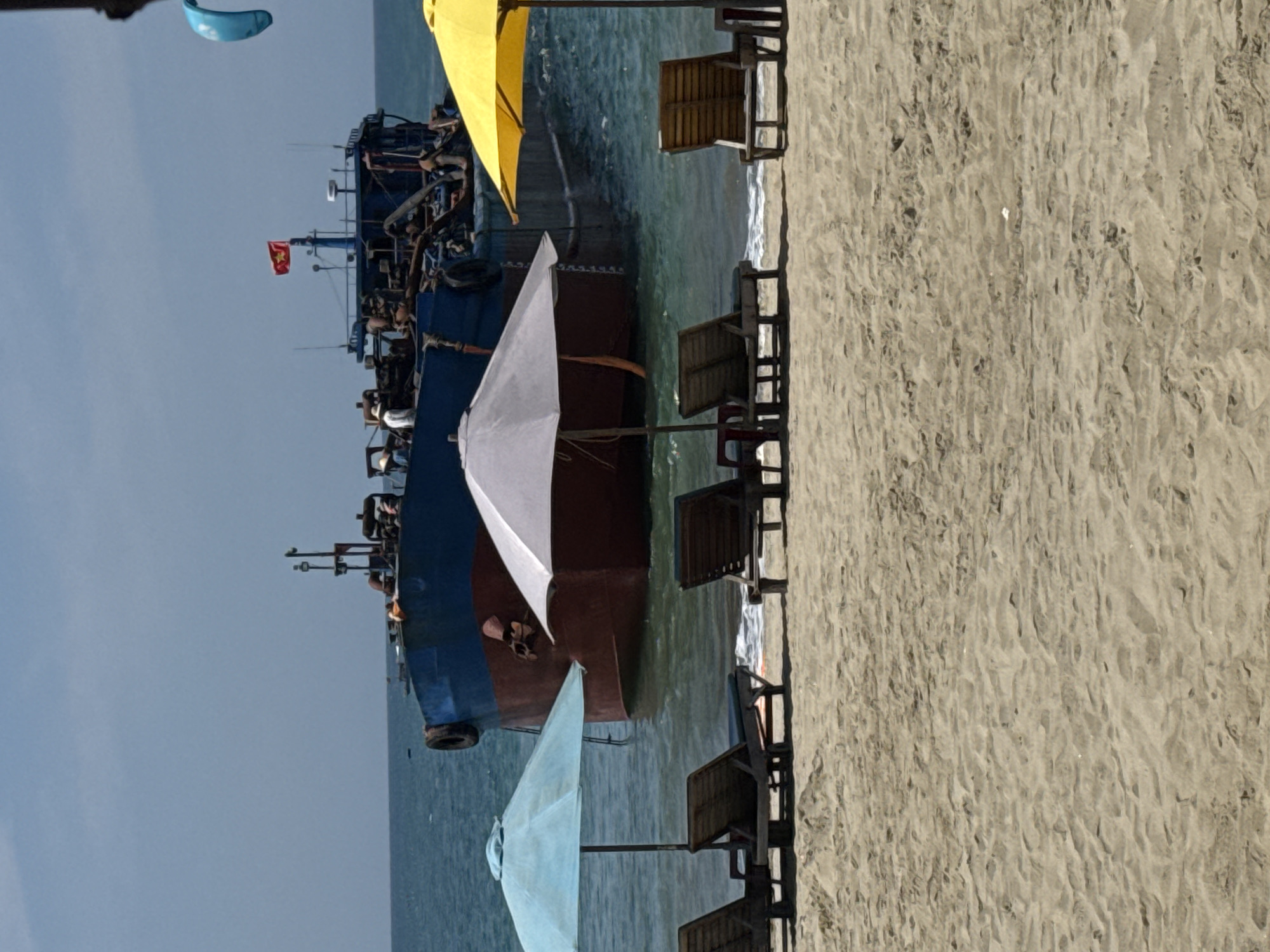 The image shows a beach scene in Đà Nẵng, Vietnam. A large blue ship with a Vietnamese flag is anchored close to the shore. Several beach umbrellas in yellow, white, and blue are present, along with empty wooden beach chairs. The beach is sandy, and the water appears calm. There are some visible people on the ship.