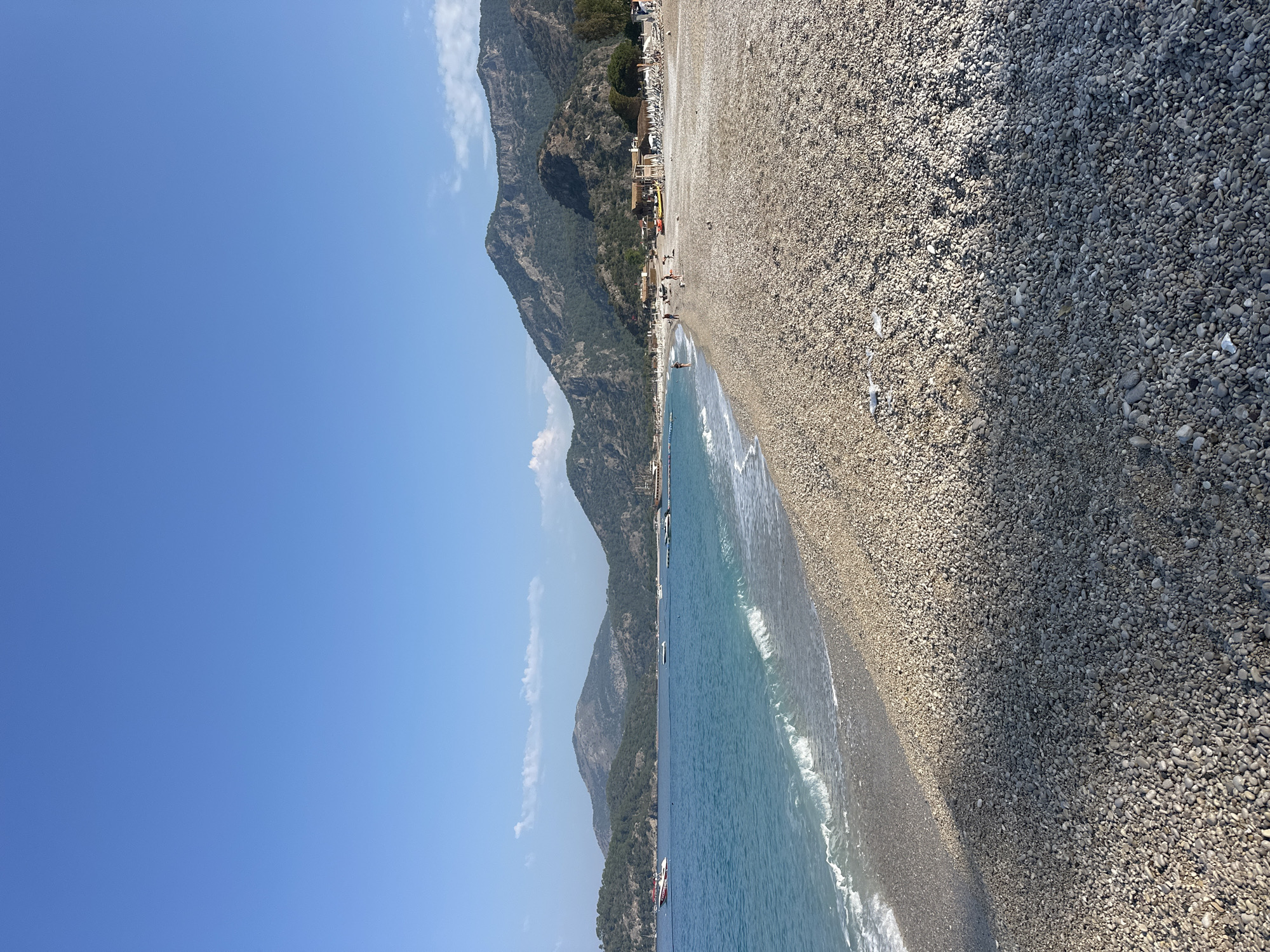 The image captures a sunny day at a pebble beach in Fethiye, Turkey, likely Ölüdeniz beach given the characteristic mountains and clear turquoise water. The foreground is dominated by a vast expanse of smooth, grey and beige pebbles, with a distinct shadow line suggesting the sun is high in the sky. Gentle waves with white foam break on the shore, creating a serene atmosphere.
In the middle ground, the clear blue water stretches towards a bay lined with lush green hills and mountains. Several boats, including a red and white speed boat and other leisure craft, are anchored or moving in the bay. Along the shoreline of the beach, there are scattered beach chairs, umbrellas, and a few buildings, indicating a developed resort area.
A small number of people are visible on the beach, some walking near the water's edge, others relaxing or engaged in activities. The sky is a brilliant, cloudless blue, completing the idyllic beach scene. The overall impression is one of a tranquil and picturesque coastal landscape.