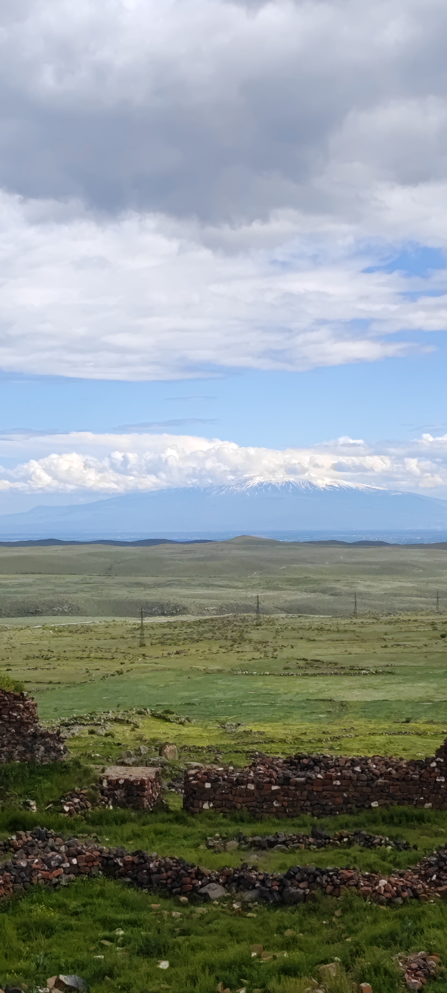 The image shows a landscape in Dashtadem, Armenia, under a sky of blue and white clouds. A snow-capped mountain is visible in the distance. A flat, green landscape stretches towards the horizon with occasional power lines. In the foreground, there are brick ruins scattered across a grassy field.