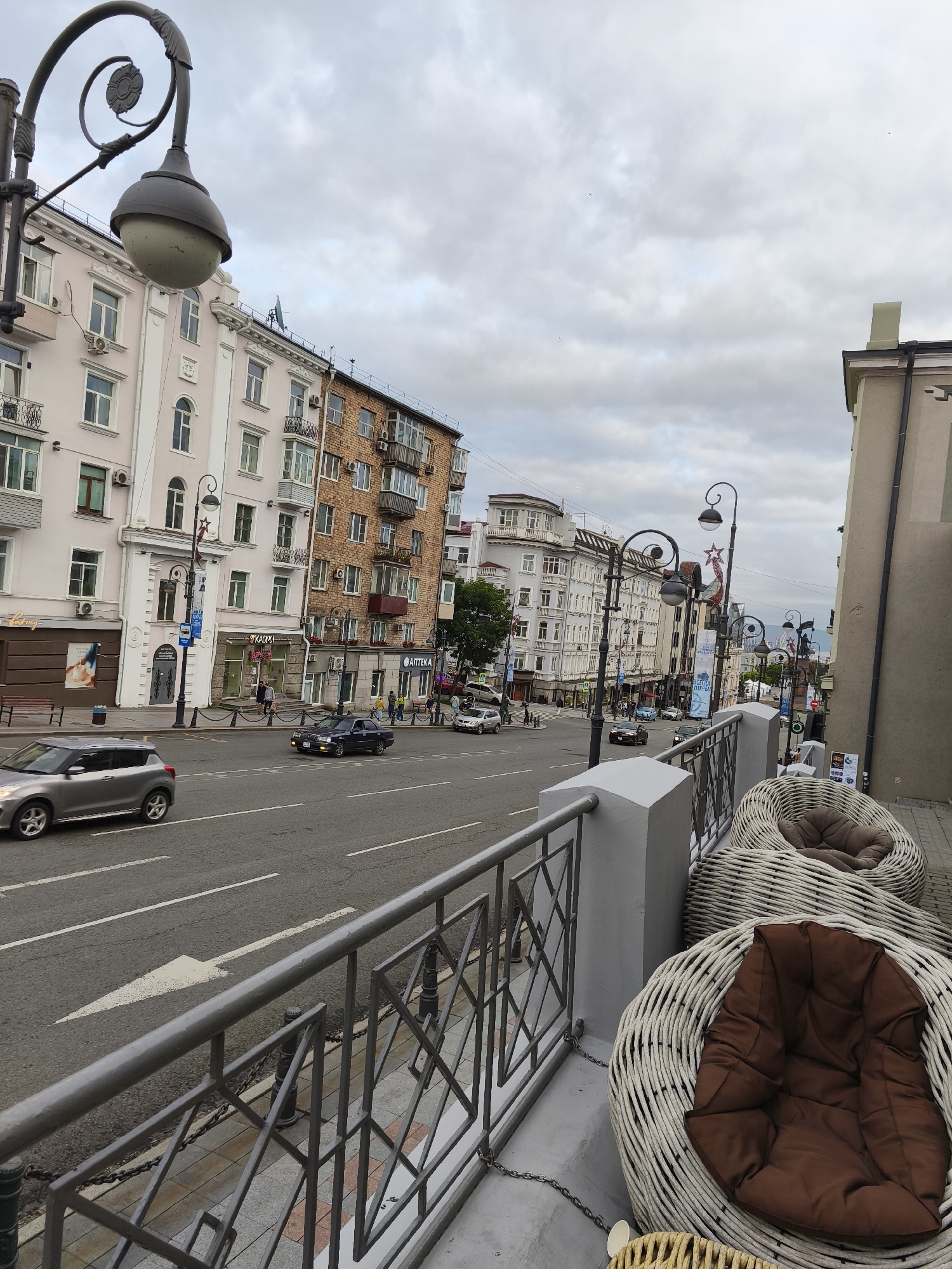 The image captures a view of a street in Vladivostok, Russia, from a slightly elevated perspective, possibly a balcony or upper-floor window. The scene depicts a street with a grey asphalt road marked with white lane dividers and a forward-pointing arrow. Several cars are present on the road, including a silver hatchback in the foreground and a dark sedan further down.
Buildings line the street on both sides. On the left, a light pink, multi-story building with classical architectural elements, including decorative moldings and arched windows, is visible. Adjacent to it is a red brick building with numerous balconies. On the right side, a tan building with some architectural detailing is partially visible.
Streetlights with ornate metalwork and globes are positioned along the sidewalk. The sidewalk features paving stones and is separated from the road by a metal railing with a geometric design.
In the foreground, on the right side, there are several large, woven wicker armchairs with brown cushions, suggesting an outdoor seating area. A metal railing separates this seating area from the street below. The sky is overcast with clouds. The overall impression is of an urban street scene on a cloudy day.