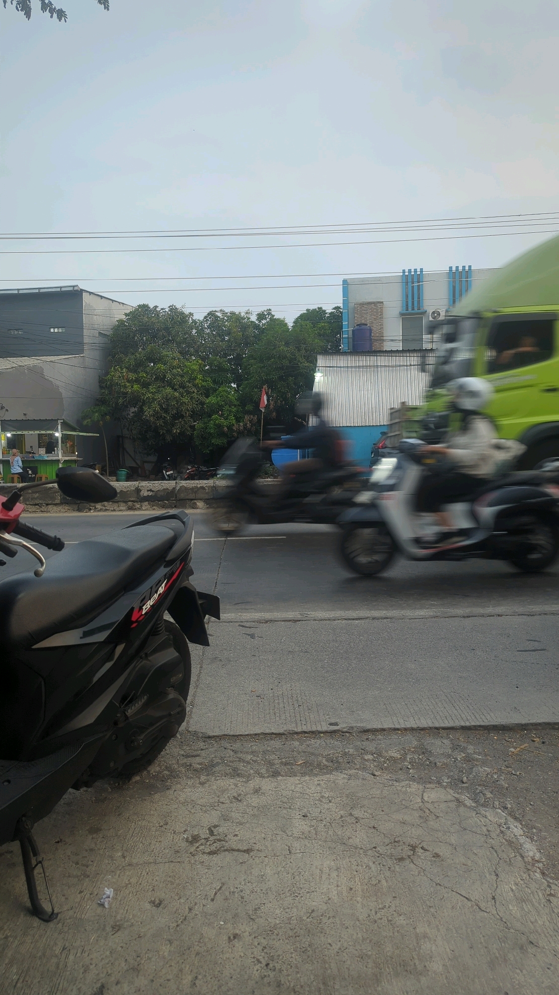 In the city of Karangtowo, Indonesia, a street scene unfolds under an overcast sky. The foreground is dominated by a black scooter, its details blurred by motion, parked on a cracked concrete sidewalk. Beyond it, a paved road shows streaks of motion blur, indicating traffic. Two motorcycles are captured mid-motion on the road: one a darker color, the other a lighter color, both with riders wearing helmets, their forms indistinct due to the speed. To the right, the rear of a bright green truck is partially visible, also blurred. In the background, a line of trees stands between the road and a row of buildings. The structures vary in design, one appearing to be a simple storefront with a green counter, another a multi-story building with white and blue accents and blue piping. Power lines are strung across the upper portion of the frame. The overall impression is a moment captured in a busy, active street environment.