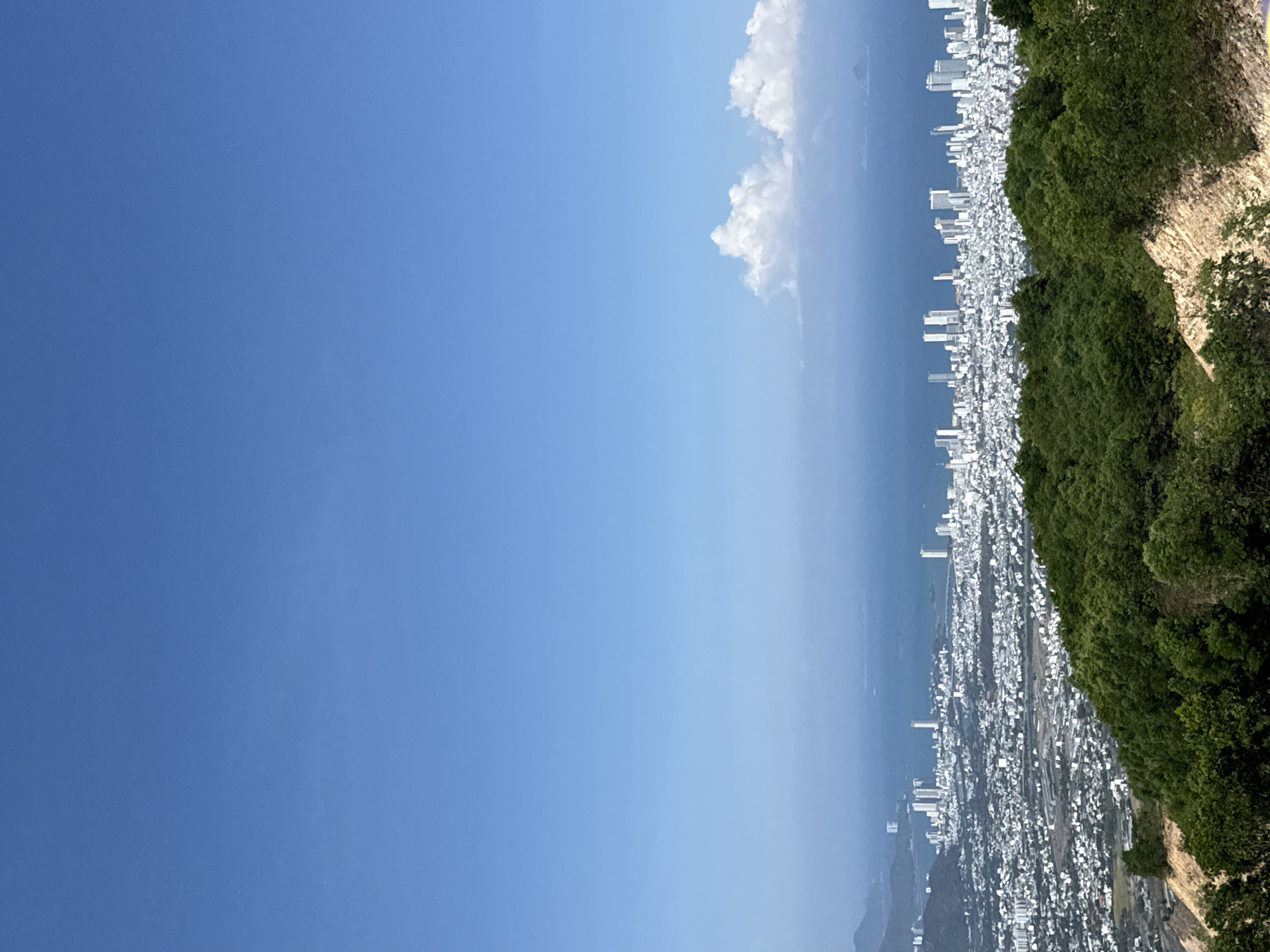 The image presents an aerial view of Nam Nha Trang Ward, Vietnam, captured on a clear, sunny day. The scene features a densely populated urban area with numerous white buildings and high-rise structures extending along the coastline. A lush green hillside borders the city. Above the city, a light blue sky is visible with a few scattered clouds. The horizon is hazy. The image is taken from an elevated position, possibly from a mountain or a high-rise building. No individuals are visible in the image.