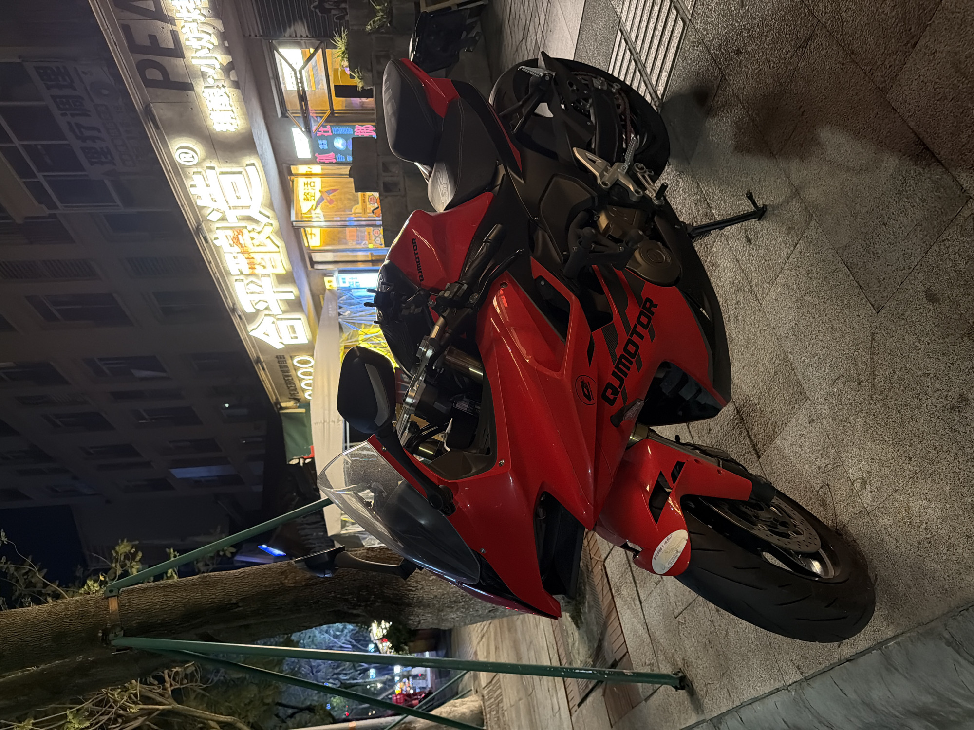 A red and black sportbike motorcycle is parked on a paved sidewalk at night in an urban environment. The motorcycle is a primary subject, showcasing its sleek design with "QJMOTOR" branding visible in black text on its red fairings. It features a clear windscreen, black seating, and its kickstand is deployed.
The setting is a vibrant city street at night, illuminated by numerous glowing storefront signs and streetlights. Buildings line the background, with windows visible against the dark sky. The sidewalk is constructed of light-colored, rectangular pavers. To the left, a thick tree trunk is supported by green metal poles. No people are visible, and the scene appears quiet despite the illuminated commercial area.
Visible text on various signs includes: "PEACEBREW 自由," "理疗调理," "合平眼造," "精酿・小炉烤," and "整活." A large number "2000" also appears on a sign. The bright lights and Chinese characters confirm the urban location, consistent with the Chongqing High-tech Zone context. The time of day is clearly night, with no specific weather conditions apparent other than a clear sky.