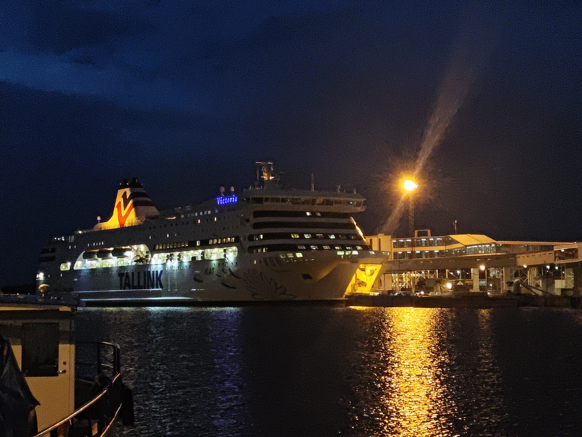 This is a nighttime, low-light photograph of the Tallink ferry "Victoria" docked at a pier in Tallinn, Estonia. The ferry, illuminated by numerous internal lights and external spotlights, dominates the frame. The "Victoria" name is clearly visible in blue neon lettering on the side of the vessel. The word "TALLINK" is also prominently displayed in large black lettering. The ship's hull is white, with a distinctive colorful pattern towards the bow.
In the background, a brightly lit terminal building or pier structure extends from the ship. A street lamp casts a strong beam of light across the dark sky and reflects brightly on the water. The water itself is dark and choppy, reflecting the lights from the ship and the terminal. The sky is dark blue with some subtle cloud formations visible.
A smaller vessel, possibly a service boat or part of the dock infrastructure, is partially visible in the foreground on the left side of the image. It is dark and its details are obscured. No people are clearly discernible in the photograph. The scene suggests a ferry terminal in operation at night, likely in the port of Tallinn.