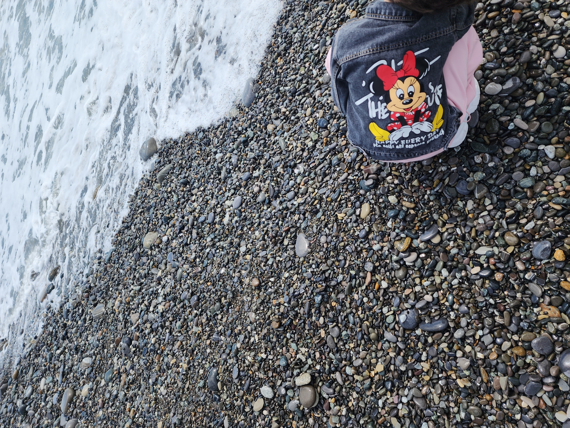 A high-angle shot shows a small child wearing a denim jacket with a Minnie Mouse graphic, seated on a pebble beach in Sochi, Russia. The ocean waves are breaking on the beach to the left, with white foam visible. The beach is made up of small, smooth, grey and brown pebbles. The child's jacket is denim with a pink lining, and the Minnie Mouse graphic is colorful. The child's head and shoulders are visible at the top of the frame. The scene is brightly lit and appears to be taken during the day.
