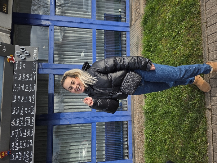 A young adult female stands centrally in an outdoor setting in Zweibrücken, Germany. She has blonde hair tied in a high ponytail and is wearing a black puffer jacket, blue denim jeans, and tan ankle boots. A dark, quilted crossbody bag with a chain strap is worn across her body. She is smiling broadly, mouth open with tongue visible, and looking directly at the camera. Her right hand is raised, holding a small, shiny, ring-like object. Behind her is a building featuring a series of blue-framed windows with vertical interior blinds visible through the glass. Above the windows, a dark sign displays multiple columns of handwritten names in white, along with text including "Lukos W 2023", "POWER", "OFF!", and "2022". A pixelated figure and an "X Y B A" graphic are also present on the sign. The foreground consists of a grey paved walkway, followed by a strip of green grass leading up to the building's base.