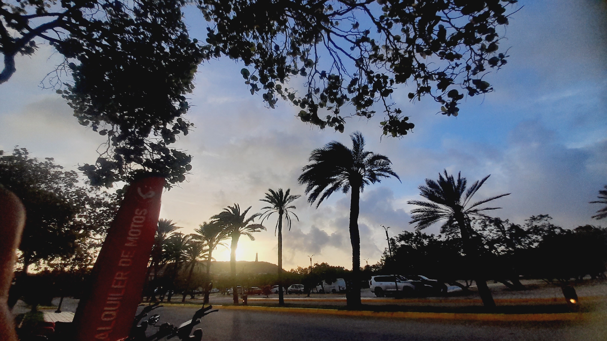 The image depicts an outdoor scene with palm trees silhouetted against a cloudy sky at dusk. A red sign with white text reading "ALQUILER DE MOTOS" is visible in the foreground, leaning against a tree. The location appears to be near a road with several vehicles, including cars and vans parked on the side. In the distance, a lighthouse or tower is visible on a hill, illuminated by the setting sun. The overall impression is a tropical or coastal environment in the city of Pedro González, Venezuela, characterized by its distinctive vegetation and a sense of twilight ambiance.