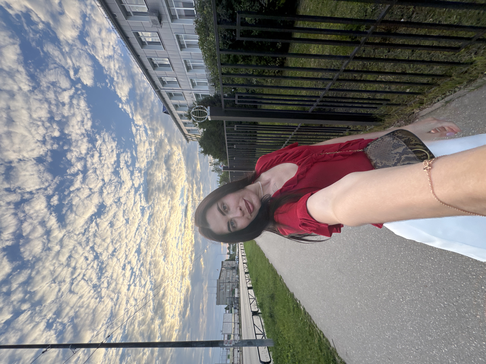 The image shows a woman taking a selfie in Chaikovsky, Russia. She is wearing a red top and a snakeskin-patterned purse is visible. The setting appears to be an urban or suburban area with a building, a fence, a sidewalk, and a grassy area. The sky is filled with white, fluffy clouds against a blue background, suggesting it might be daytime with fair weather. The woman is looking at the camera with a slight smile. There is a thin necklace visible on her neck. The image is oriented sideways.
