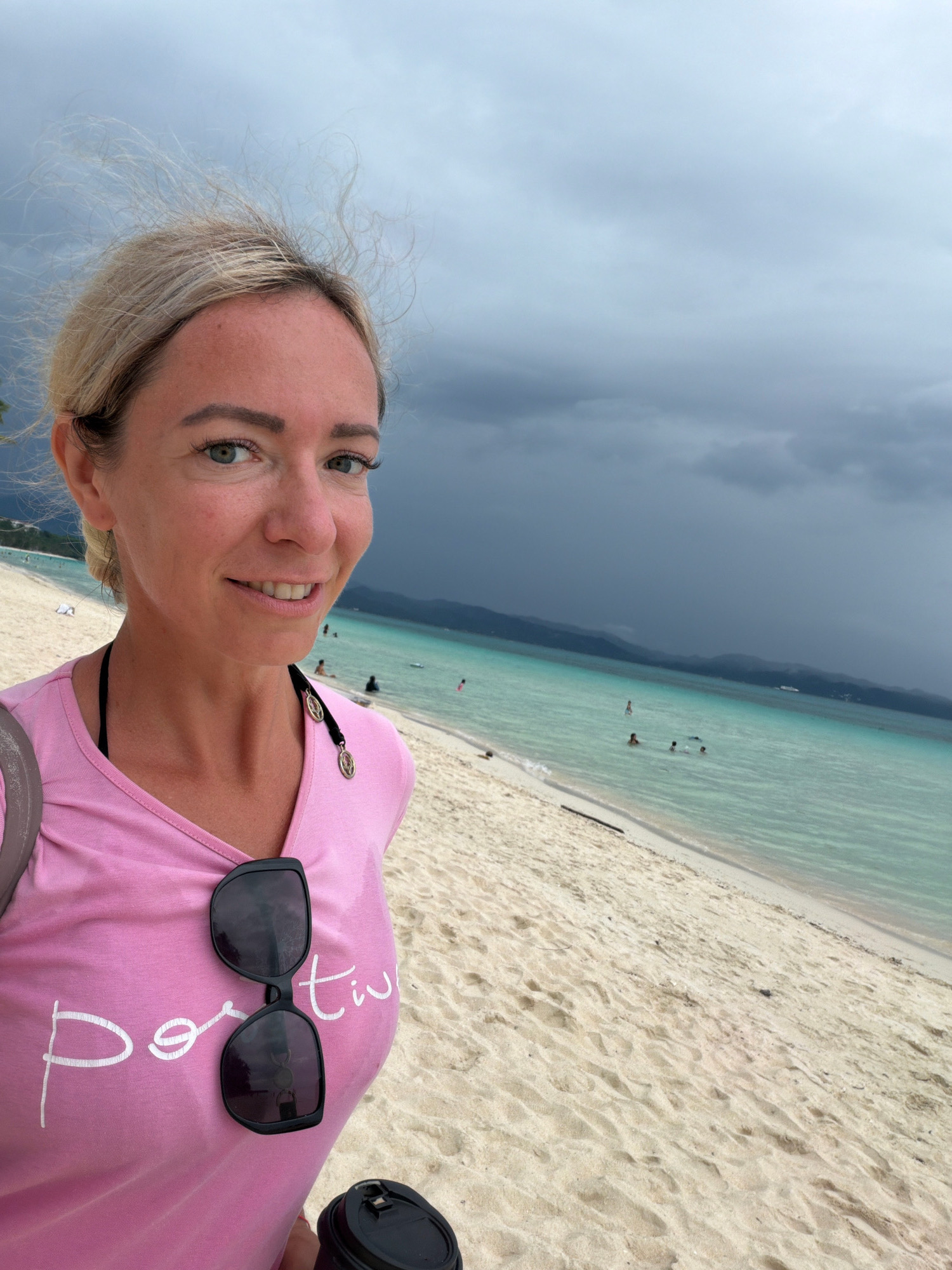A woman with blonde hair pulled back is taking a selfie on a sandy beach. She is wearing a pink t-shirt with white cursive writing that says "positive" and a black bikini top. Sunglasses are resting on her t-shirt, and she is holding a black coffee cup. In the background, the turquoise water of the ocean meets a sandy shore, with a few people swimming in the distance. The sky is overcast with dark storm clouds forming over the horizon, casting a dramatic atmosphere. The location is identified as Malay, Philippines, likely on Boracay Island given the typical scenery of white sand beaches and clear turquoise waters often associated with the area, even under stormy skies.