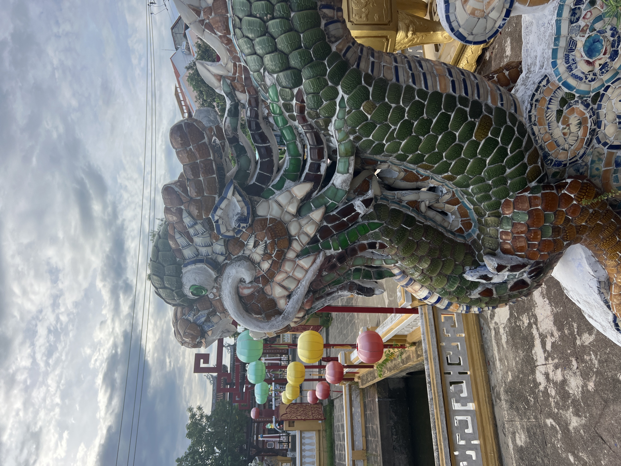 In Hoi An, Vietnam, a mosaic dragon statue is prominently featured in the foreground, its head and scales detailed with colorful ceramic fragments. Behind the statue, a pathway lined with colorful, spherical lanterns in shades of green, yellow, and pink leads across a narrow bridge. The bridge spans a body of water and is flanked by yellow and white railings with decorative cutouts. In the background, traditional Vietnamese architecture with red accents can be seen, along with trees and buildings. The sky above is overcast with dramatic clouds, and power lines are visible. The scene appears to be an outdoor public space, possibly a temple or a garden, given the cultural elements present.