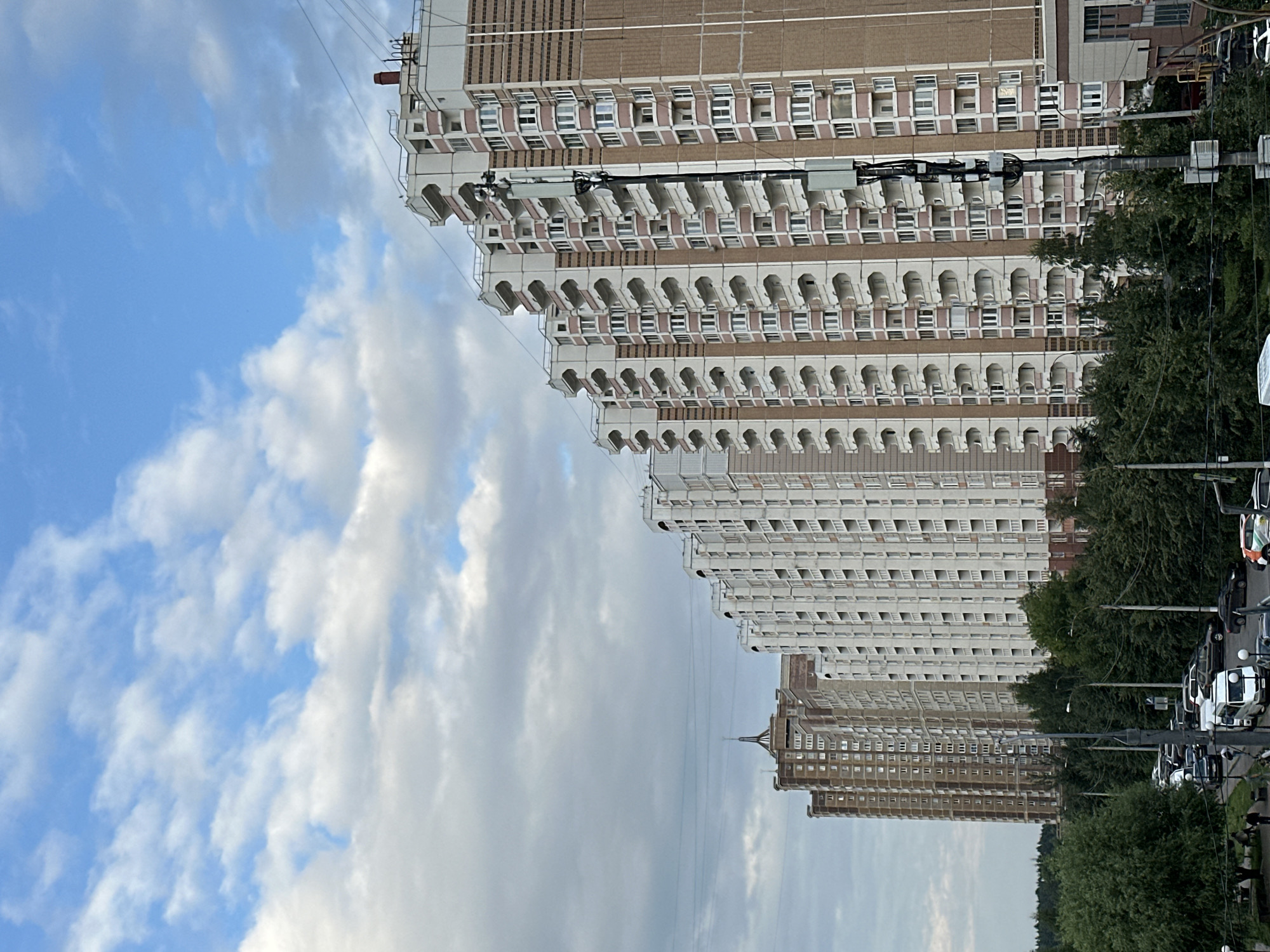 The image captures a view of apartment buildings in the city of Podolsk, Russia, under a partly cloudy sky. The buildings are tall, multi-story structures with a repeating pattern of balconies and windows. They appear to be made of brick and concrete, with beige and pink accents on the facade. In the foreground, there is a line of trees and a street with several cars parked and in motion. A few individuals are visible walking on the sidewalk. Electrical wires are strung between poles and across the buildings, and a cellular antenna tower is mounted on a pole in front of one of the buildings. The overall impression is one of a residential urban environment.