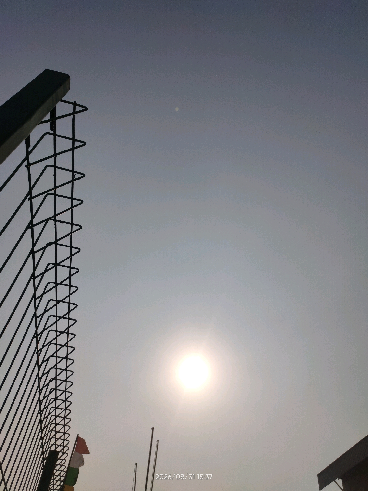 The image depicts a bright, hazy sky with the sun prominently displayed near the bottom center. A black metal fence with a distinctive zigzag pattern dominates the left side of the frame. In the lower left quadrant, a small Indonesian flag, appearing red and white, is visible, along with other indistinct colorful flags. The date and time stamp "2026/08/31 15:37" is overlaid on the bottom right of the image. The scene is likely outdoors, possibly in an urban or semi-urban environment given the presence of the fence and flags, though no specific landmarks identifying Telagaasih, Indonesia are clearly discernible. The overall atmosphere is one of a bright, possibly warm day with a haziness obscuring distant details.