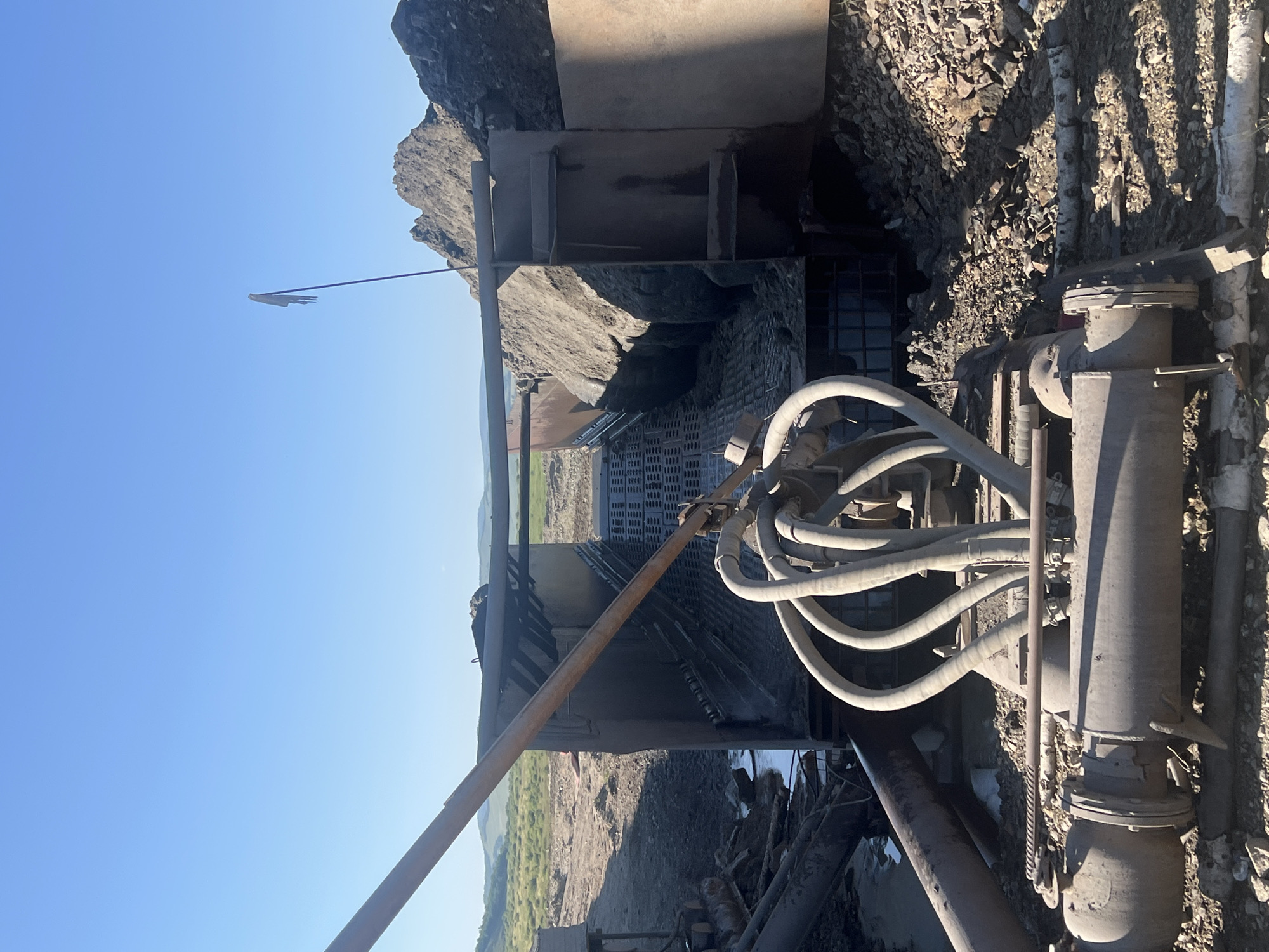The image shows a view of industrial equipment in an outdoor setting, possibly a mining or construction site. In the foreground, there are large, thick pipes with various couplings and fittings, some appearing weathered and rusted. Several flexible, ribbed hoses are connected to a central point, curving upwards. Behind this, a large metal structure, likely a screening or sorting machine, is visible. This structure has a grated surface and appears to be filled with gravel and dirt. Further in the background, piles of dark earth and rocks are present, contrasting with a green, hilly landscape under a clear blue sky. A tall, thin pole with a tattered flag at its top stands against the sky to the right of the frame. The overall impression is one of an active, though possibly unkempt, industrial operation in a rural or undeveloped area. There is no indication of the city of Kozlovo, Russia in the image.